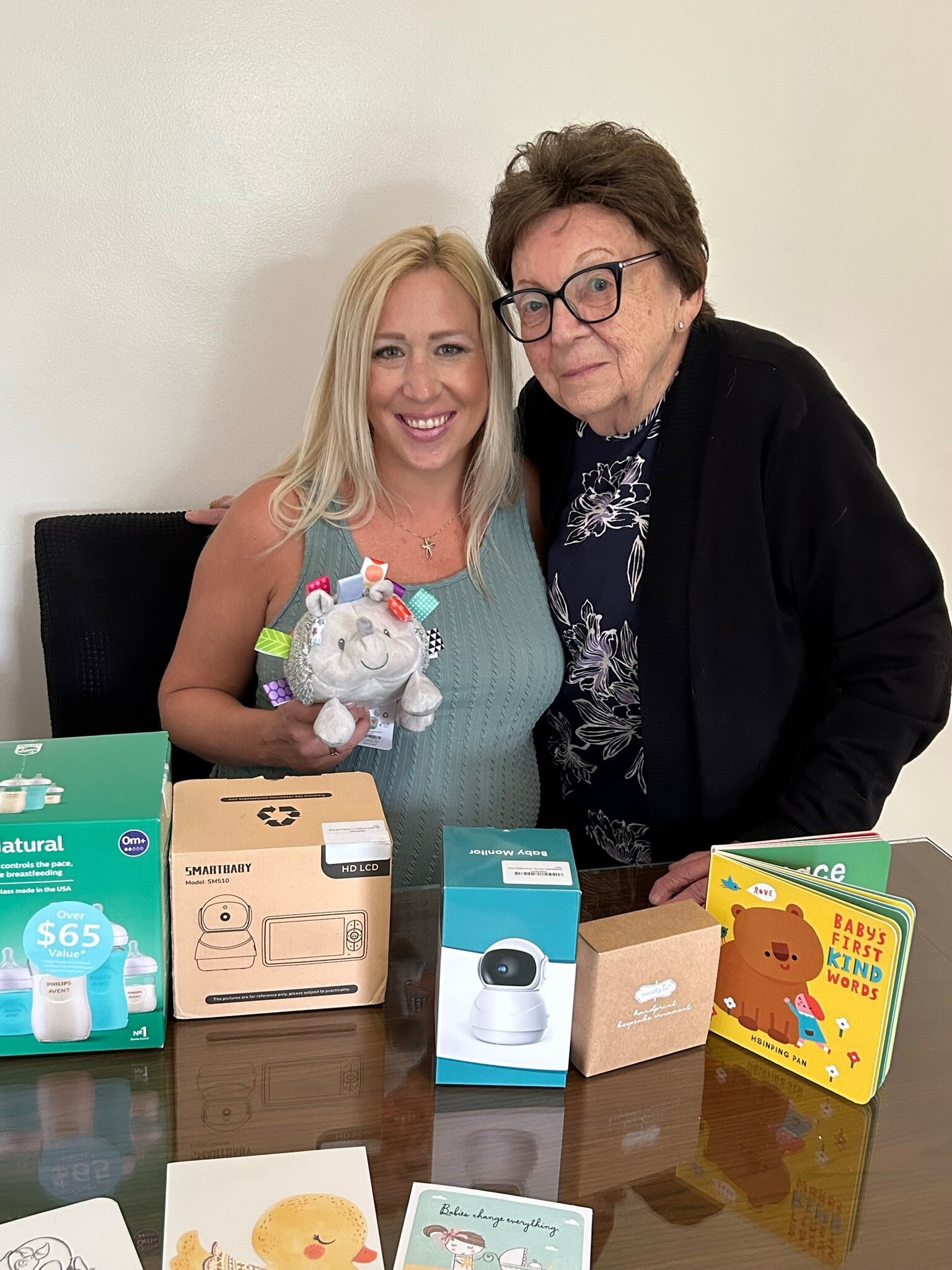 Two women pose for a photo behind a table filled with baby and toddler product boxes, smiling at the camera, one holding a plush toy.