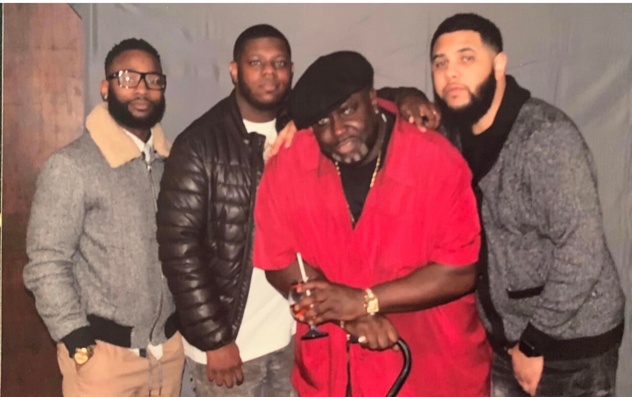 Four men posing together indoors; center man in a red shirt holds a drink and cane, flanked by three friends in jackets.