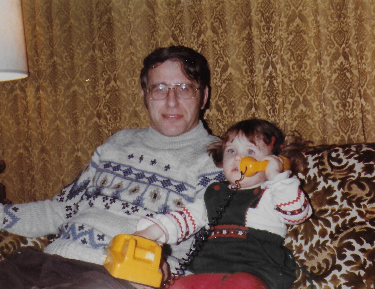 Man wearing glasses sits on a patterned sofa with a young girl holding a yellow toy phone to her ear.