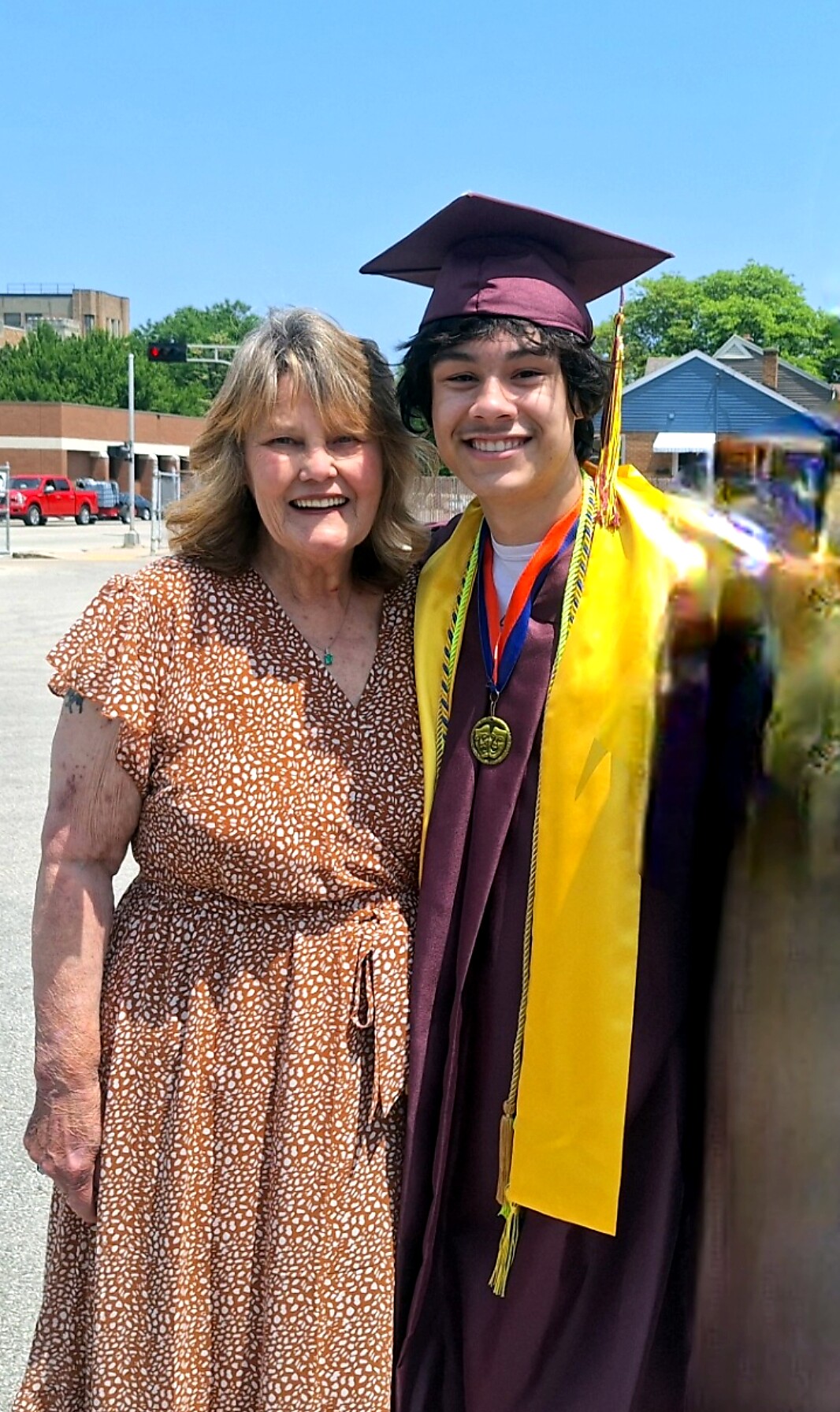 Young graduate in a maroon cap and gown with a gold stole, smiling beside an older woman outdoors.