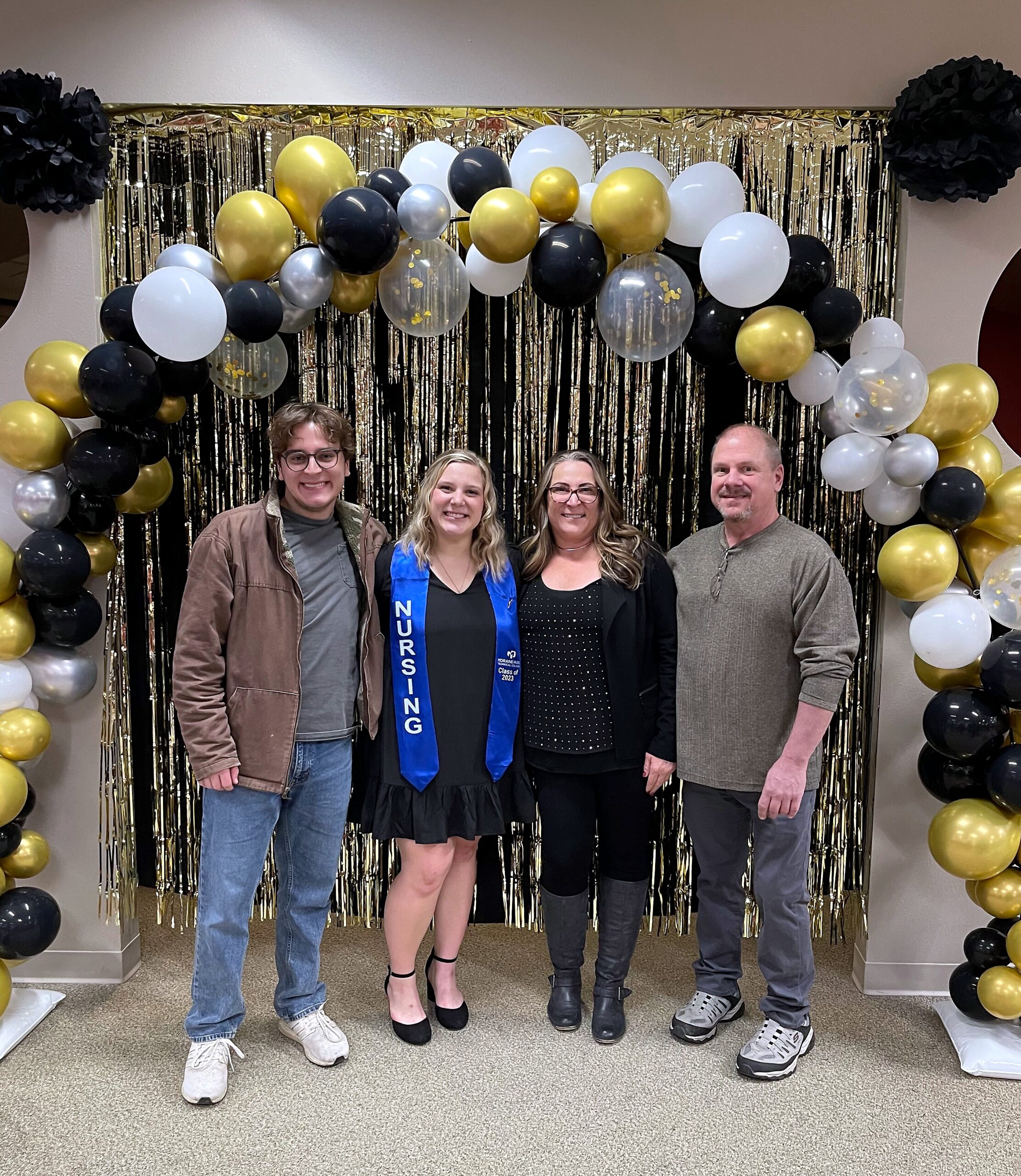 A group of four adults posing in front of a gold, black, and white balloon arch indoors; center woman wears a blue nursing sash.