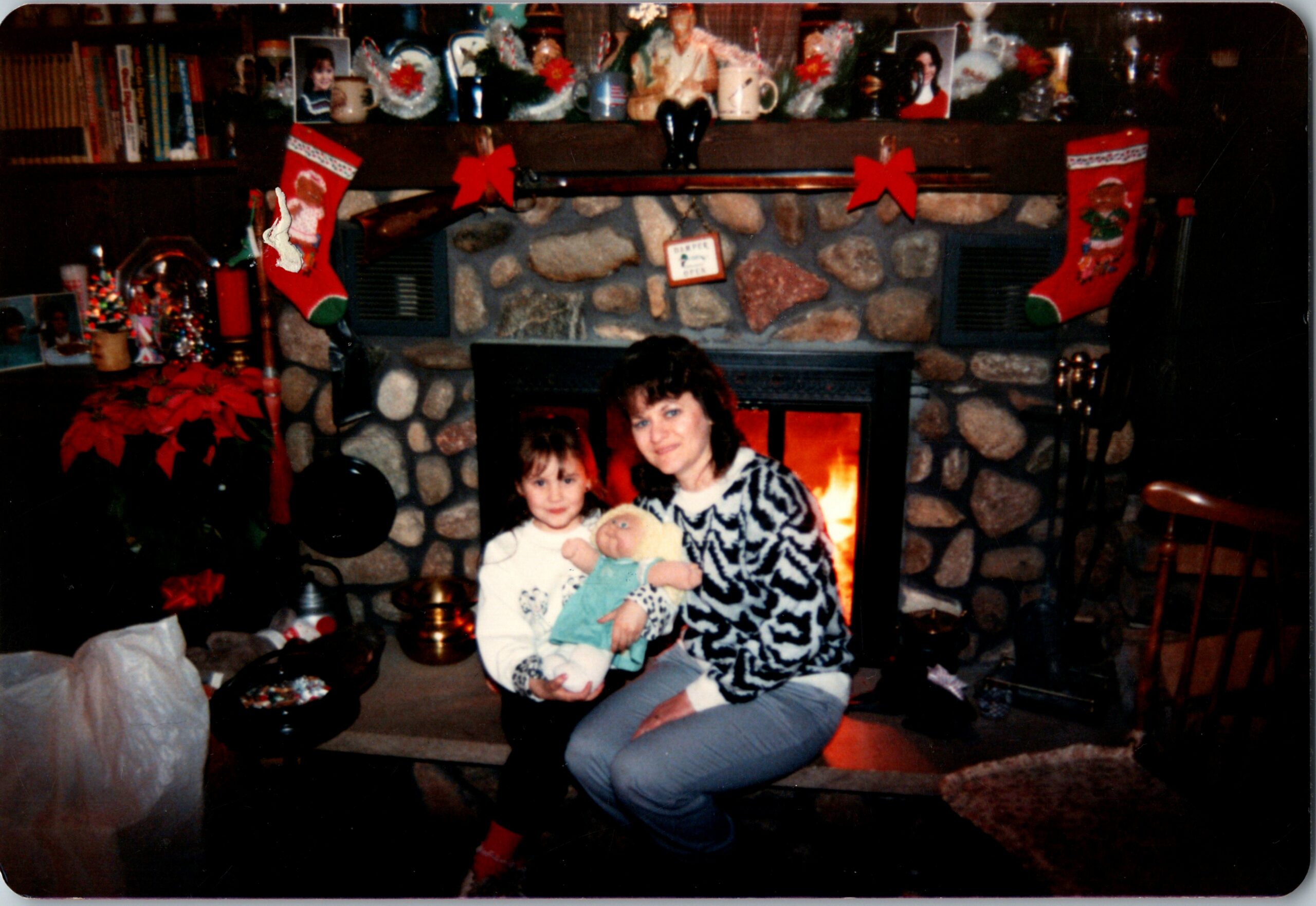 Woman in a patterned sweater sits with a child by a stone fireplace, holding a doll, Christmas decorations around them.