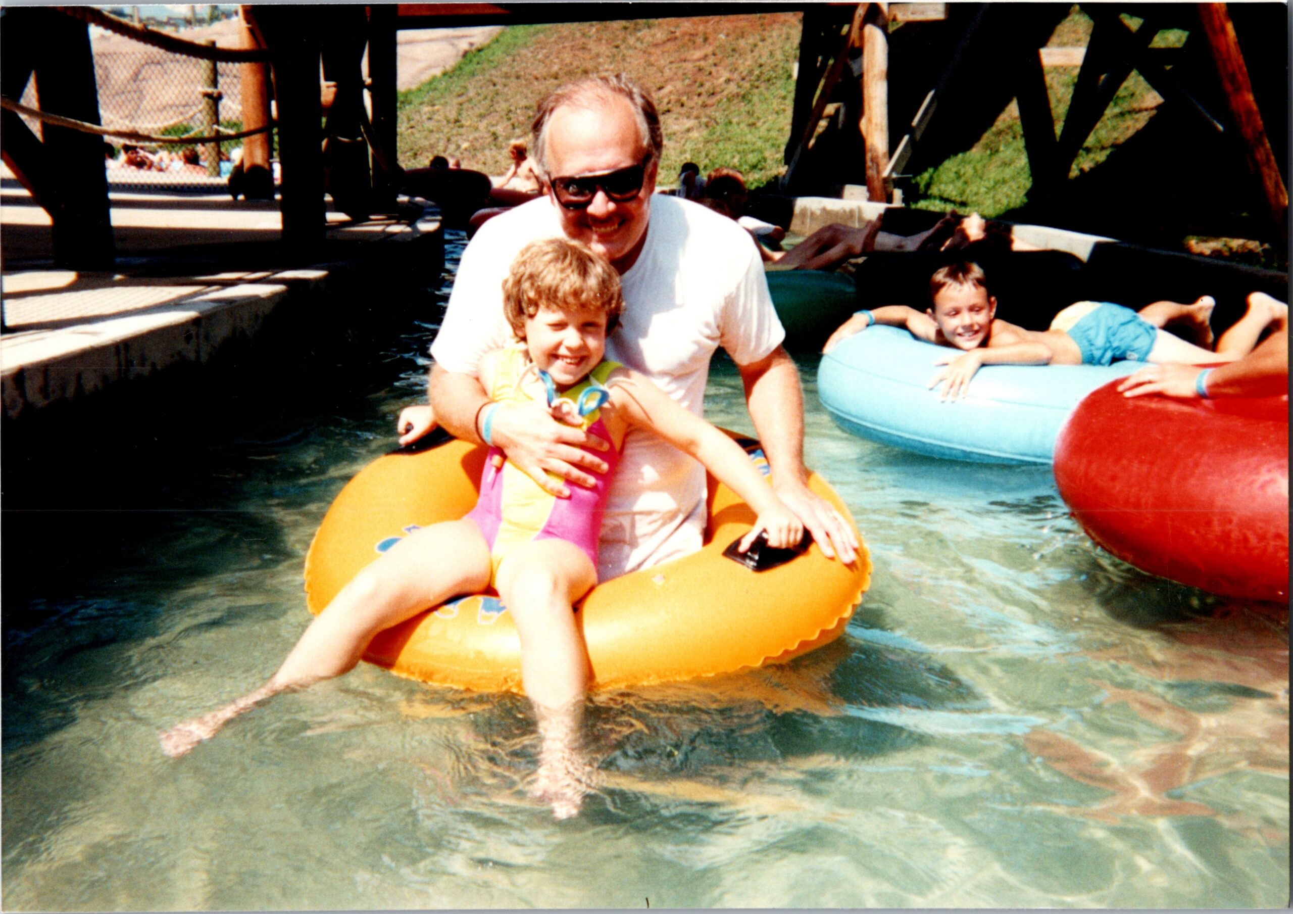 Man wearing sunglasses helping a smiling child in a yellow life vest on an orange inflatable tube in a water park pool.