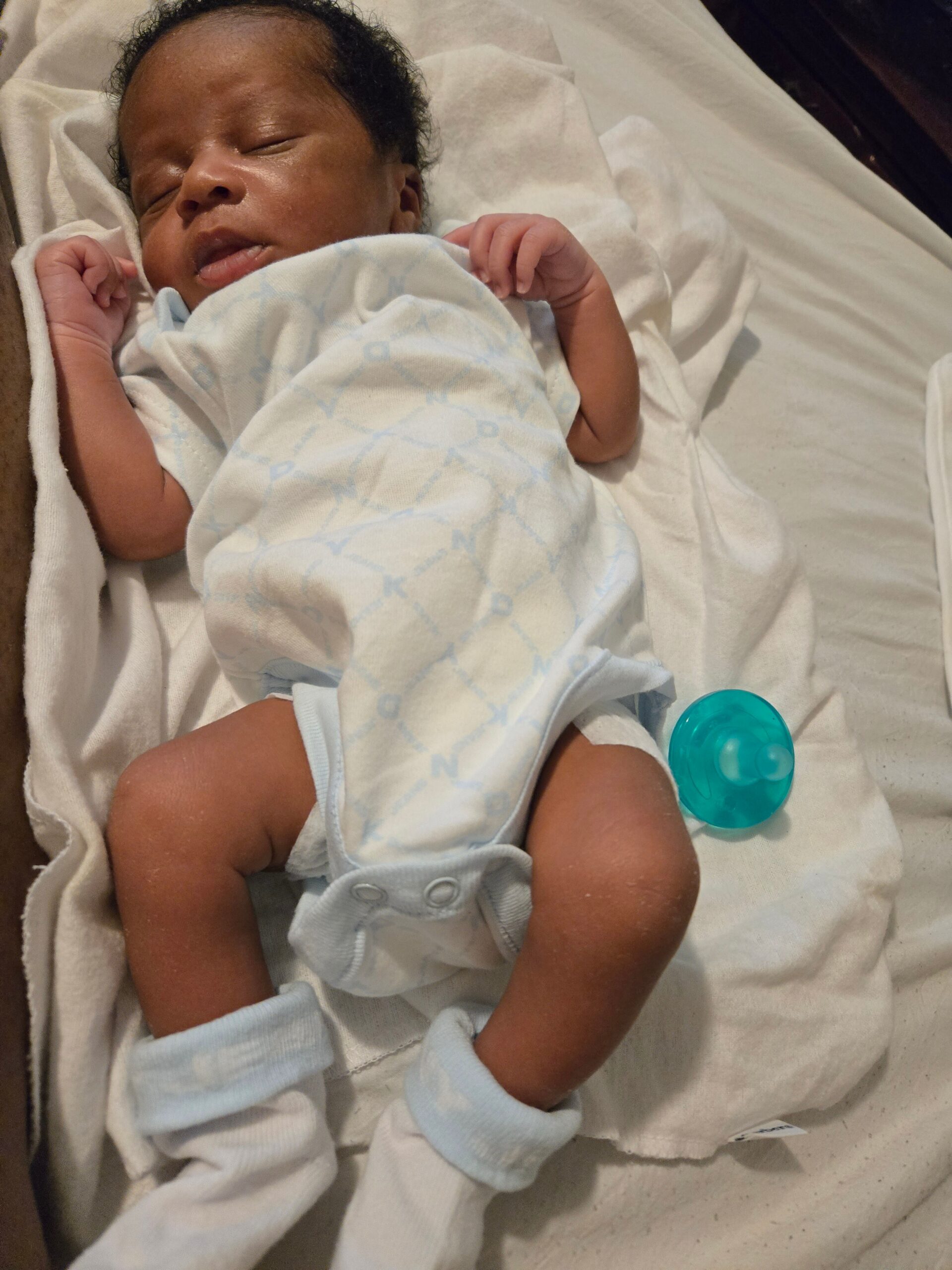 Sleeping newborn baby swaddled in a light blue blanket on white sheets, with a blue pacifier nearby