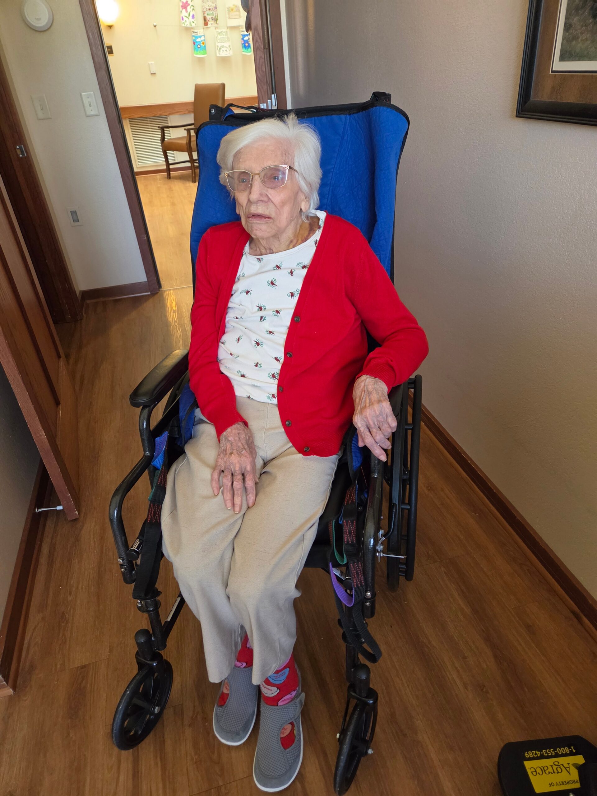 Older woman with white hair and glasses, wearing a red cardigan and light clothing, sits in a blue wheelchair in a hallway.