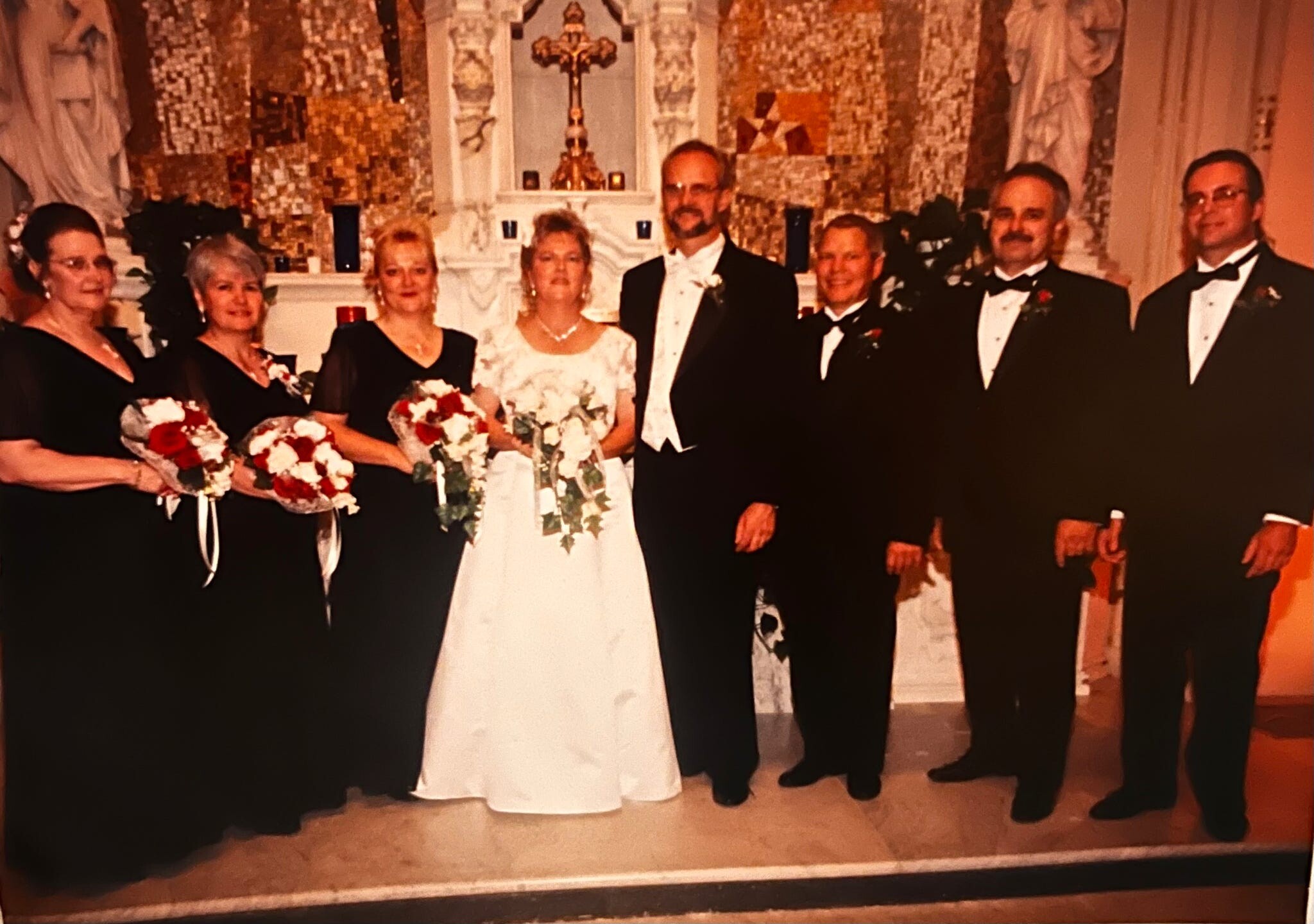 Bride in white gown with bouquet posing with wedding party in black-tie outfits in a church altar setting.