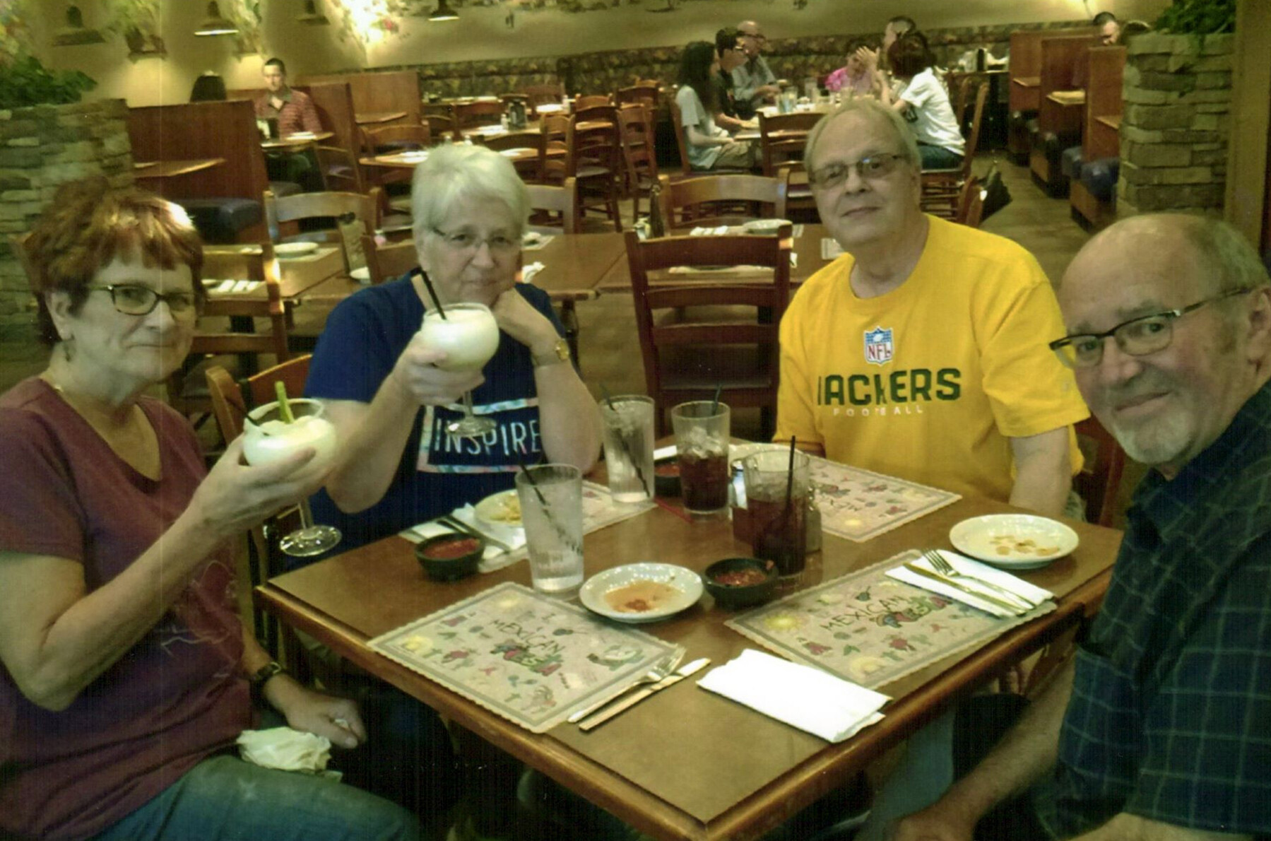 Four adults sit around a restaurant table, toasting with milkshakes and drinks while smiling at the camera.