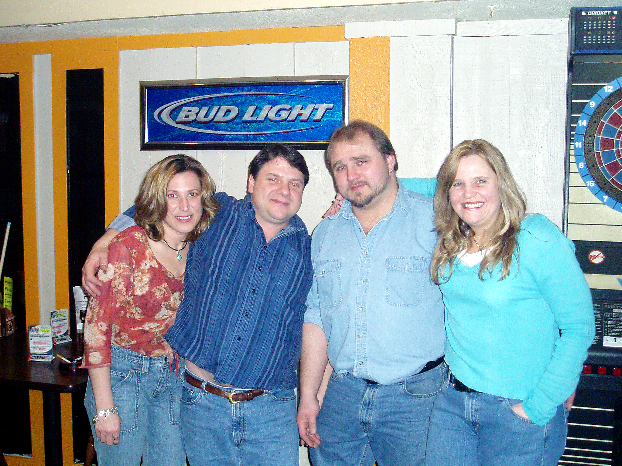 Four adults posing together at a bar, with a Bud Light sign and a dartboard visible in the background, smiling at the camera.