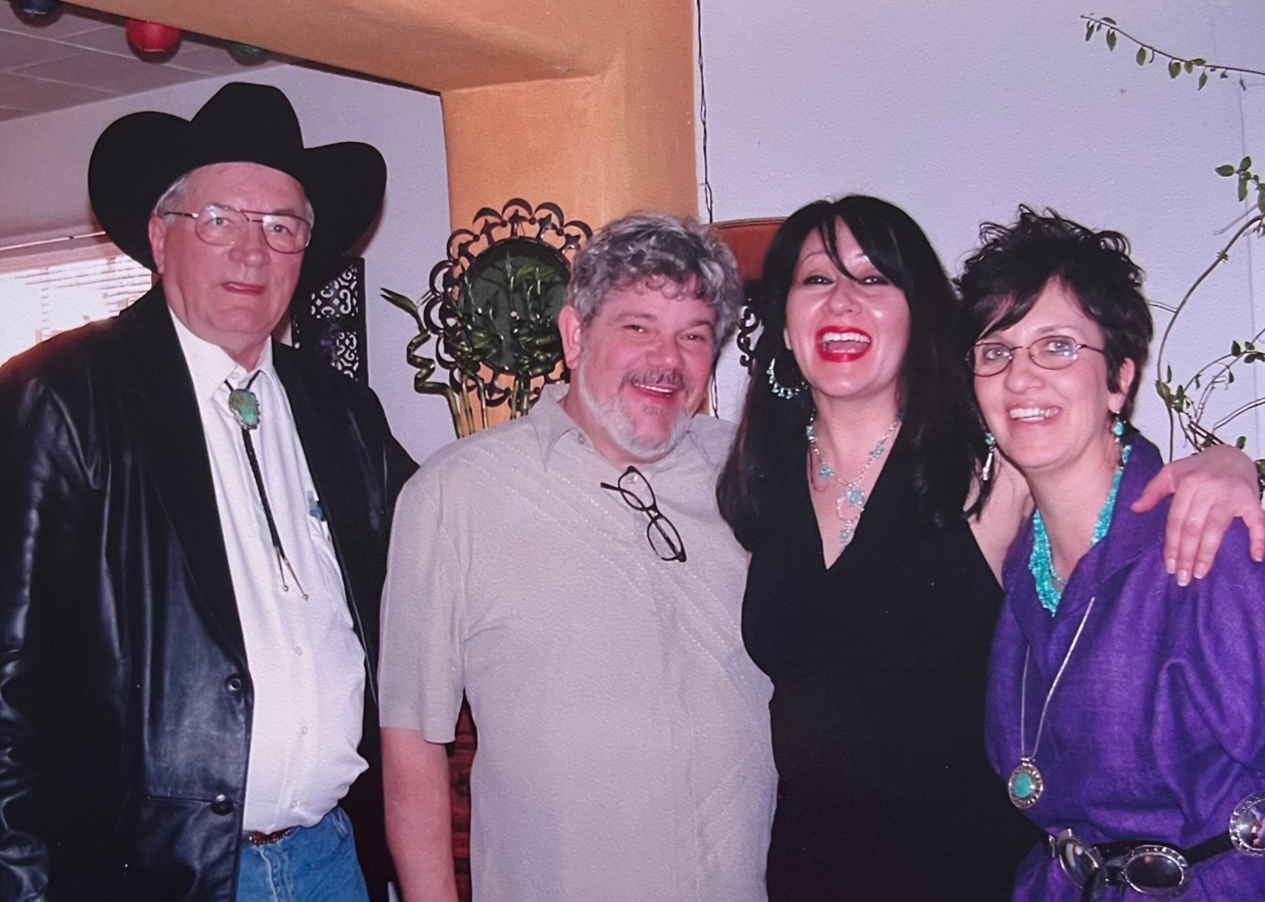 Four adults posing together indoors at a party, smiling for the photo; man in cowboy hat and leather jacket on the left, other three beside him wearing casual to semi-formal outfits.