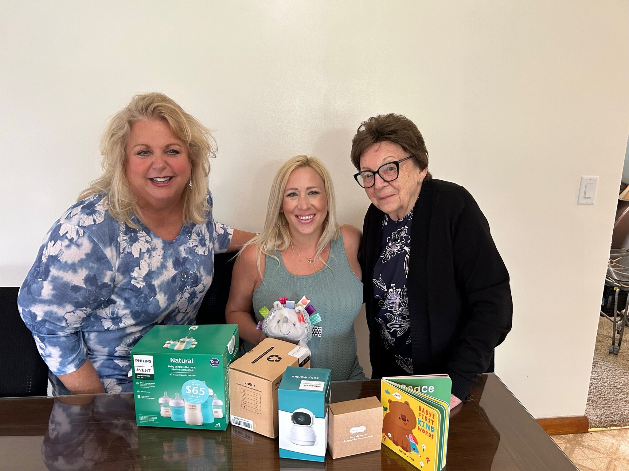 Three women smile at a table displaying assorted baby products and boxes.