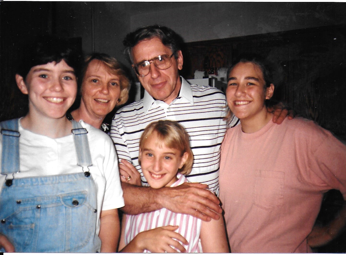Five people smiling and posing for a casual group photo indoors, with two young women on either side, an older man wearing glasses in the center, and a younger girl in front.