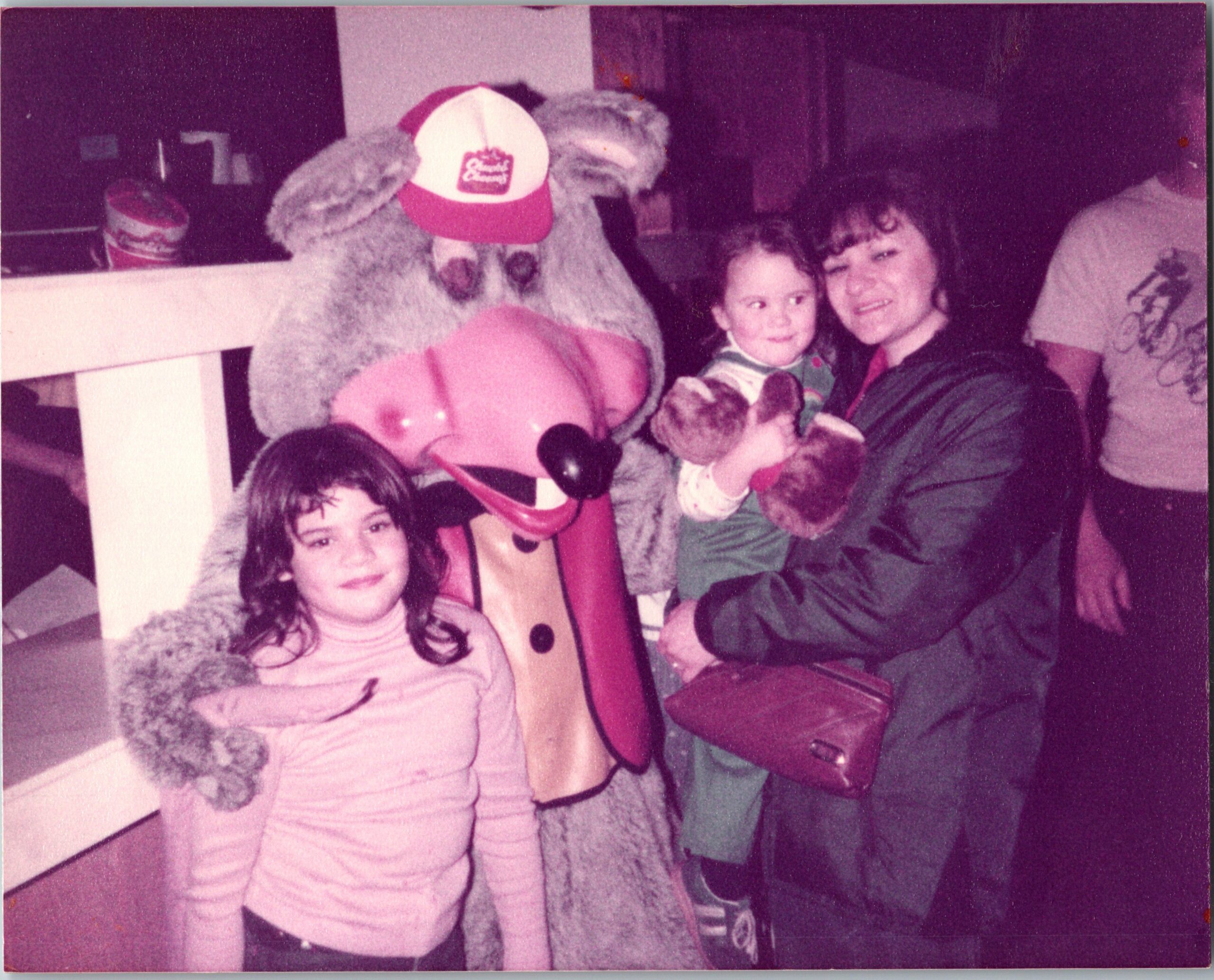 Child in pink striped outfit posing with a large gray furry costume character wearing a red cap at a party or event.