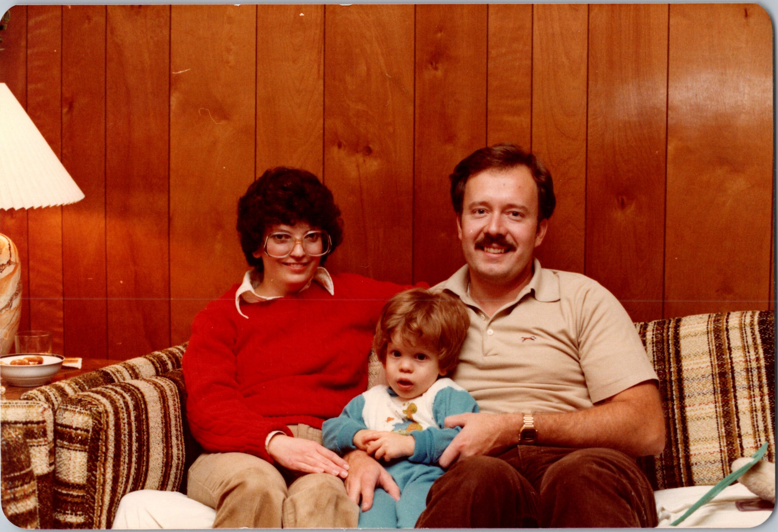 Family portrait on a plaid sofa in a wood-paneled room: woman with glasses in red sweater, man with mustache next to a young child wearing blue, all smiling.