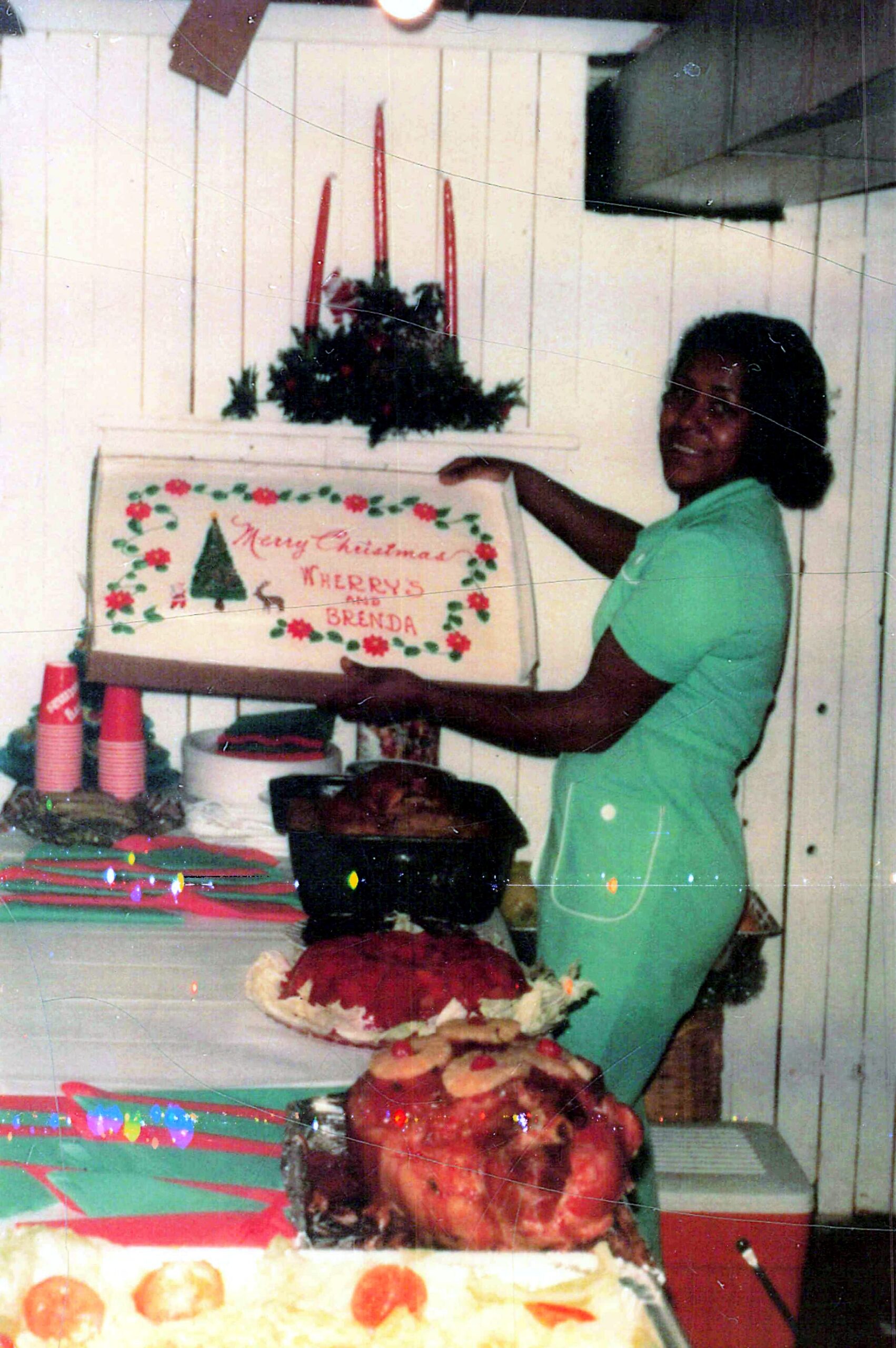 Woman in a green shirt smiles as she presents a large decorated Christmas cake with red candles and greenery; a sign reads 'Merry Christmas' in the background.