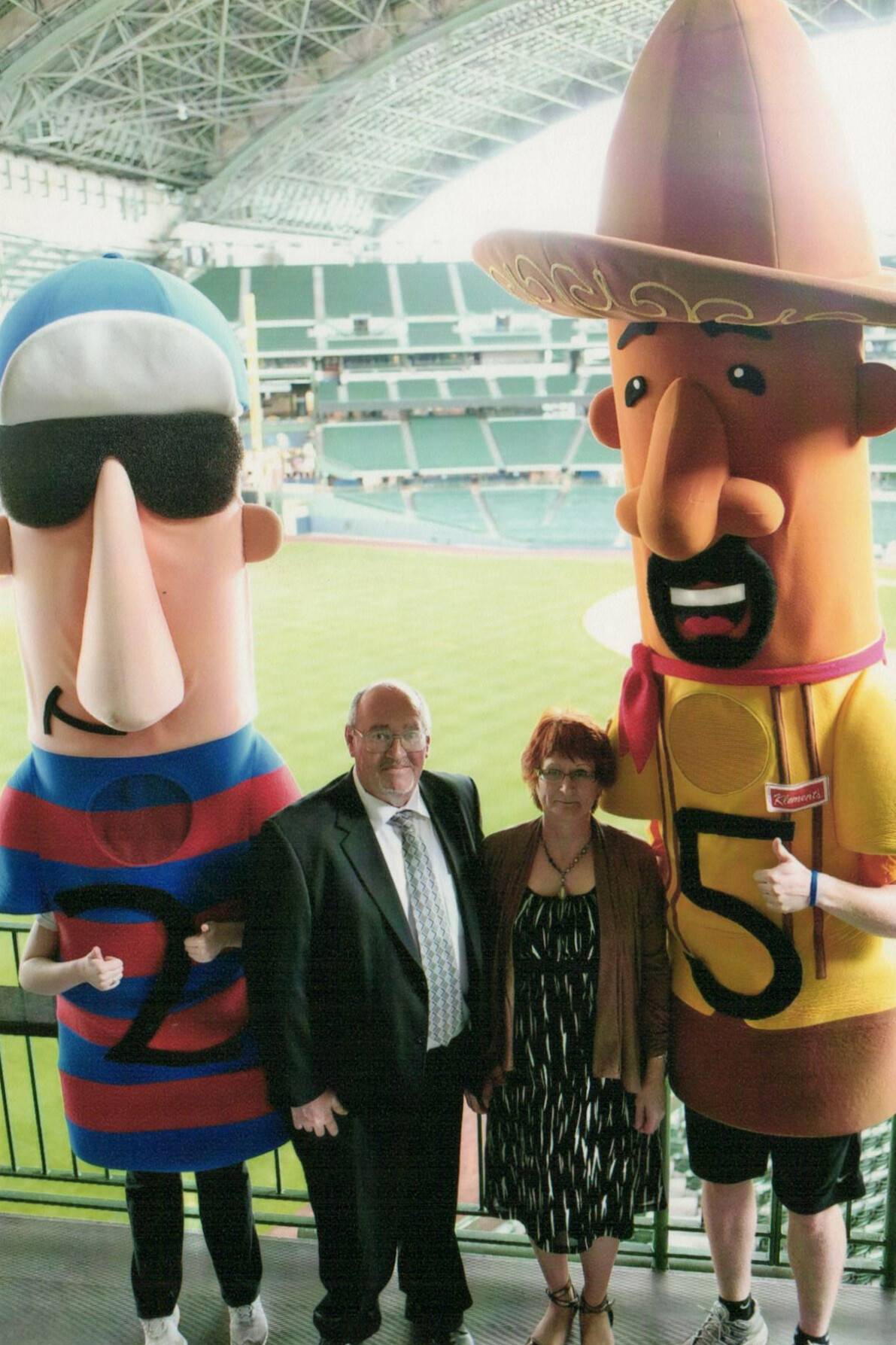 Man and woman stand with two large cartoon mascots at a baseball stadium concourse, posing for the photo.