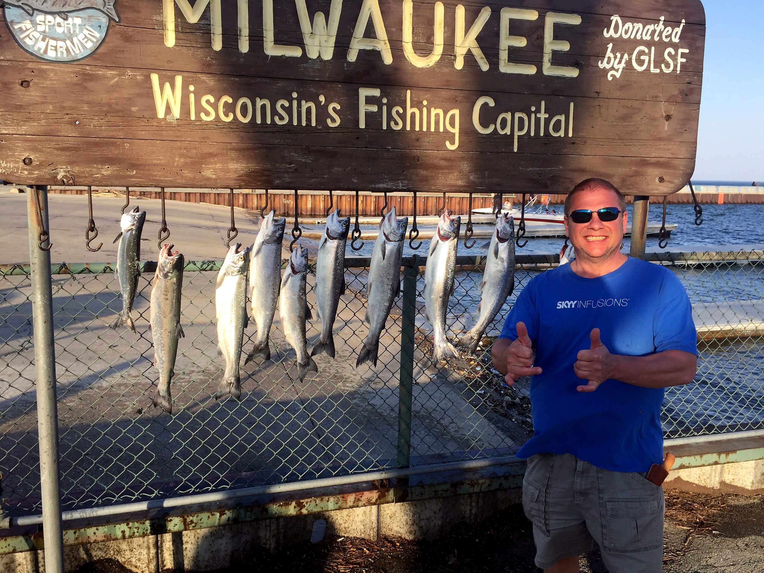 Wooden sign reading Milwaukee's Fishing Capital with fish hanging on hooks and a man in sunglasses giving thumbs up.