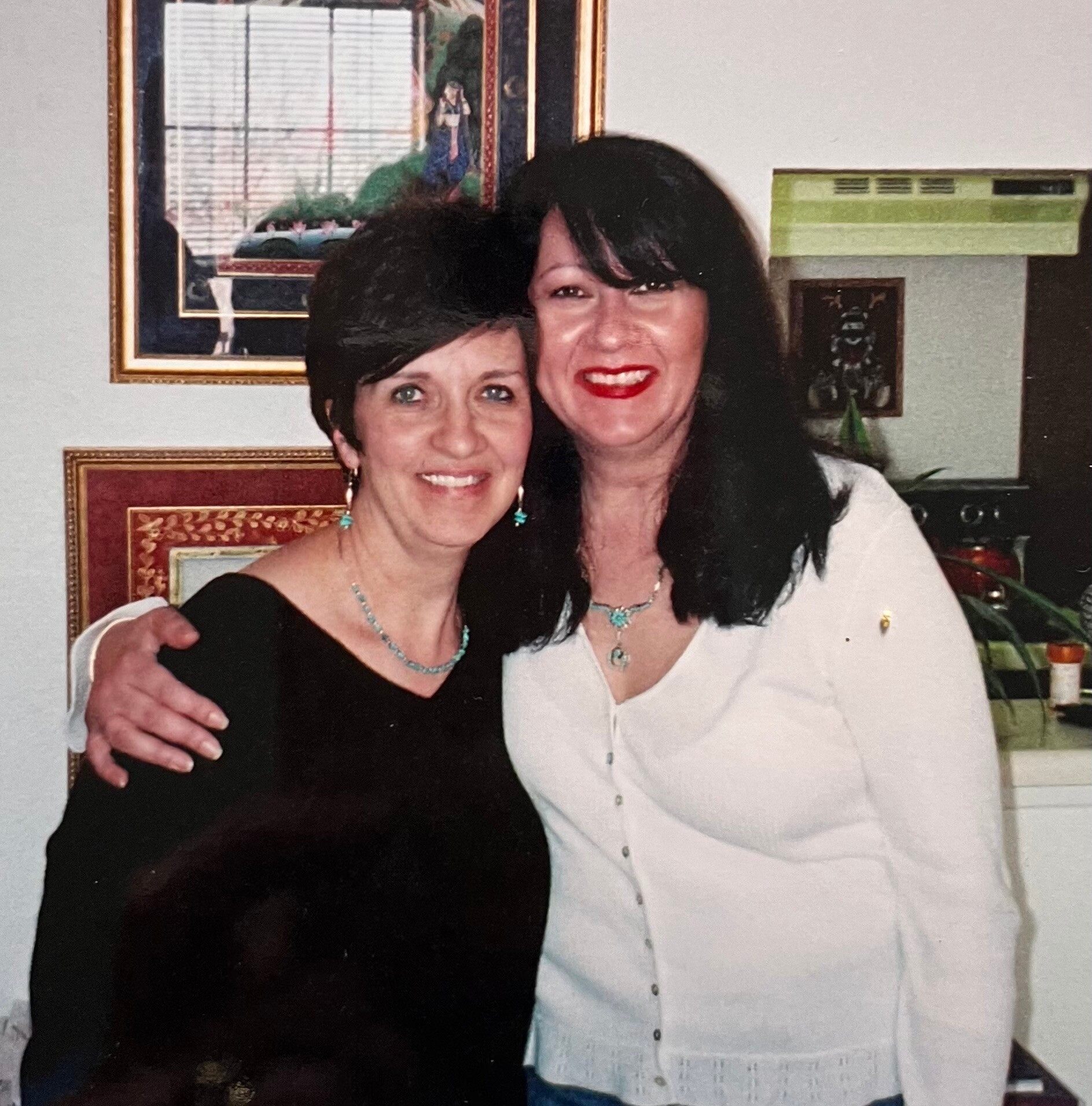 Two women smile and pose together indoors, one in a black outfit with turquoise jewelry and the other in a white blouse, framed art and a green box behind them.