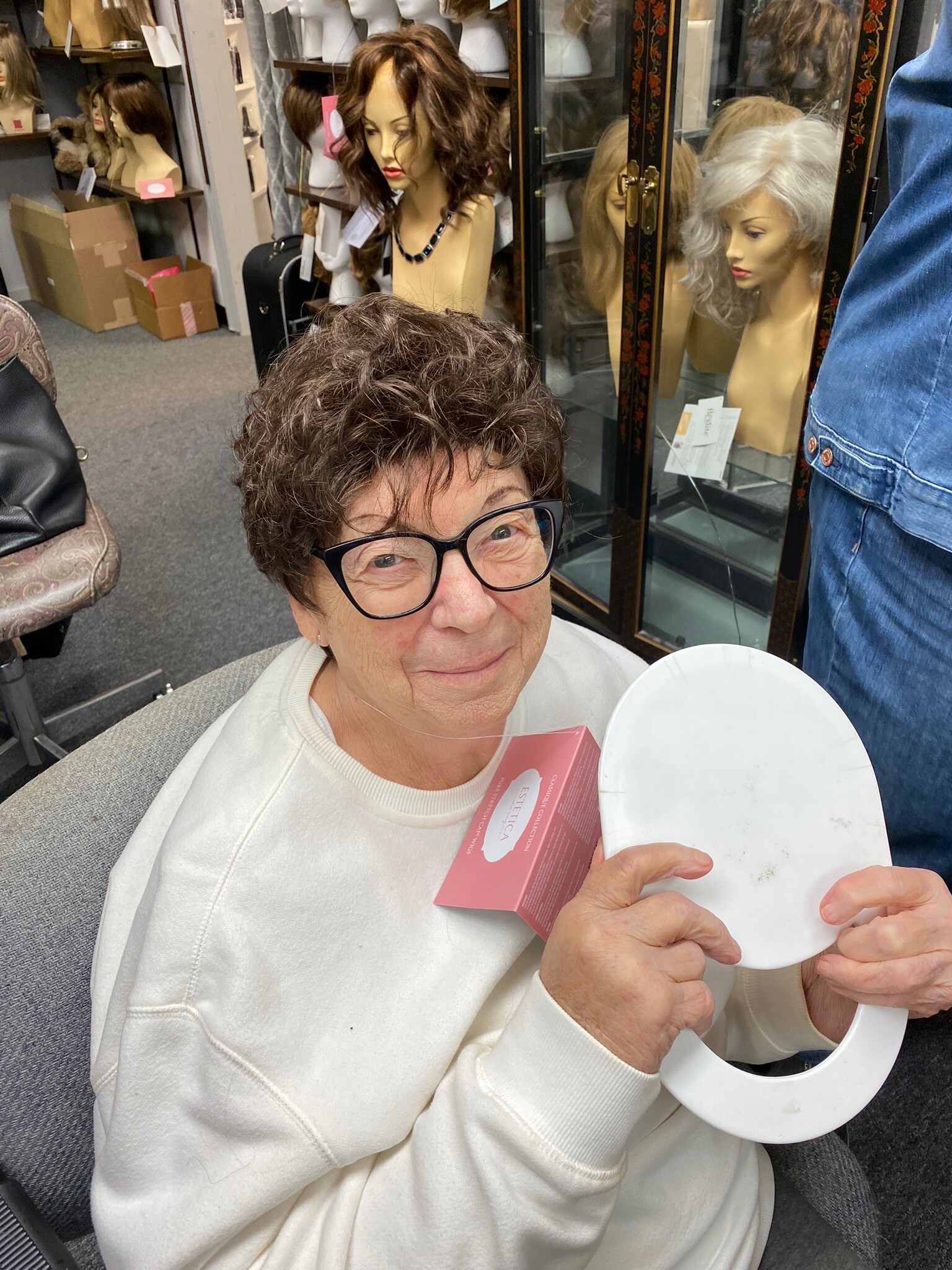 Older woman with short curly hair and glasses sits in a wig shop or salon, holding a pink product box and a white plate/plastic tray.