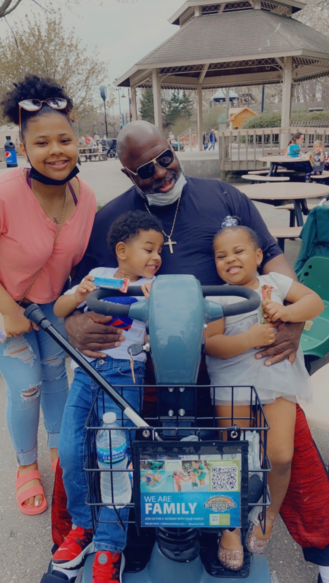 Family of four posing together on a blue scooter in a park; father wears sunglasses and a cross necklace, mother beside them, two kids in front with snacks or treats in hand.