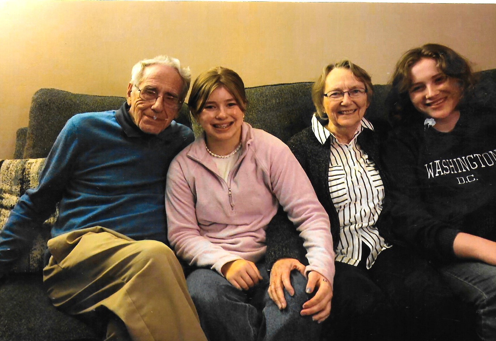 Four people sit closely on a dark sofa, smiling at the camera: an older man in a blue shirt, a teenage girl in a pink jacket, an older woman in a striped blouse, and a young person in a Washington DC hoodie on the right.