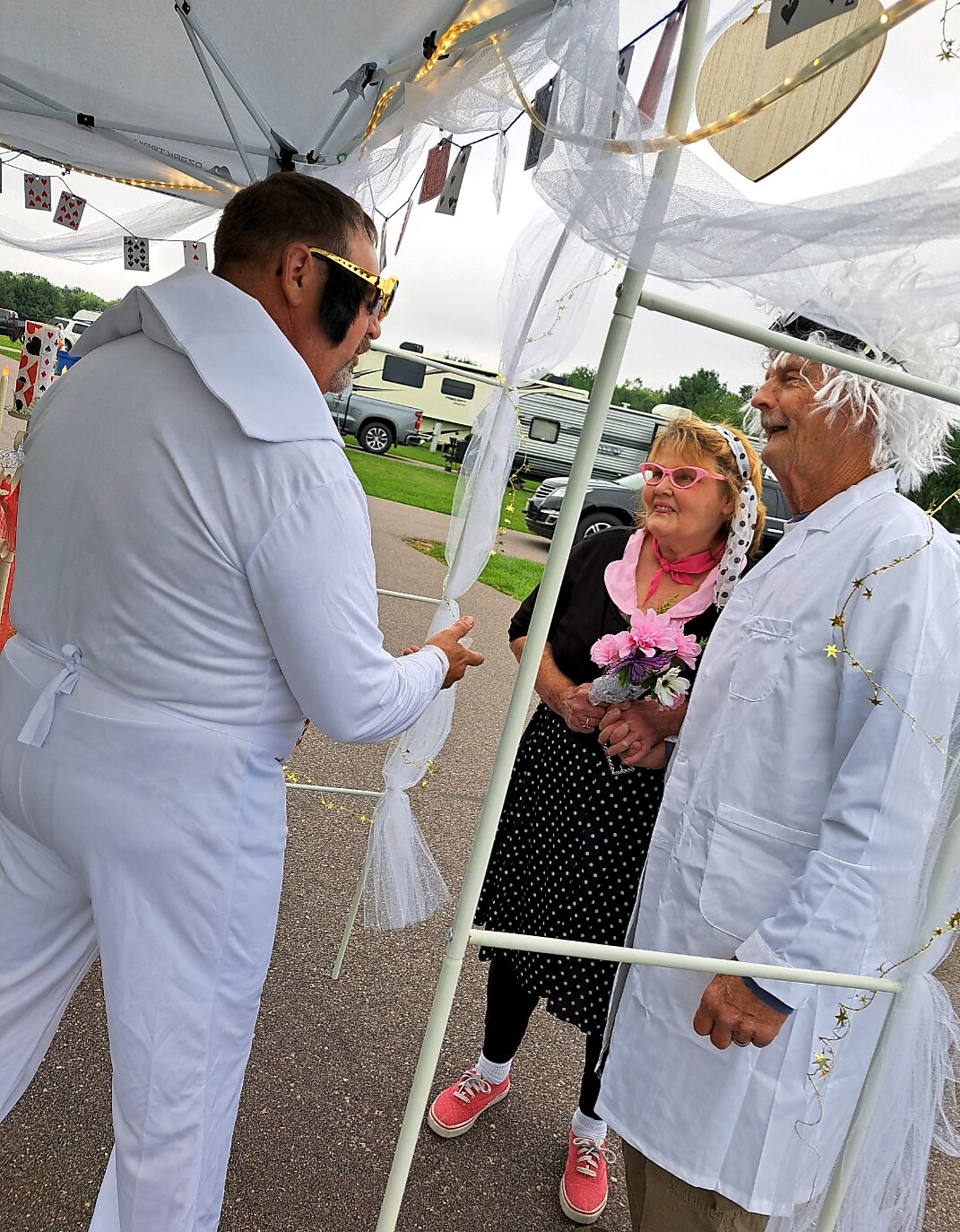 Three people in costume under a decorated white canopy at an outdoor event; man in a white Elvis-like suit talks to a smiling couple holding flowers in festive attire.