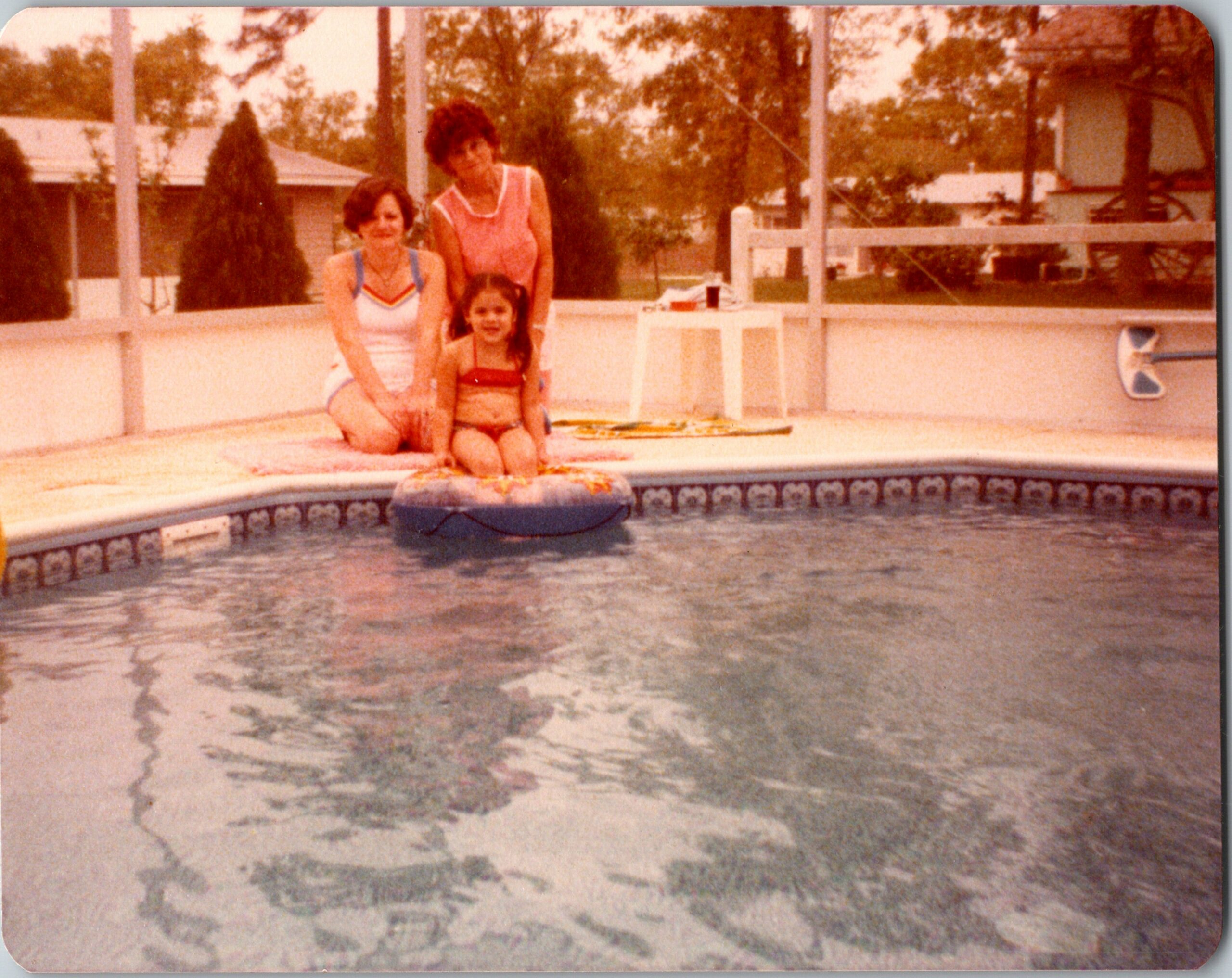 Girl sits on a colorful pool float at the edge of a backyard pool, with two women kneeling beside her as the water and a fence surround the scene (warm, faded photo).
