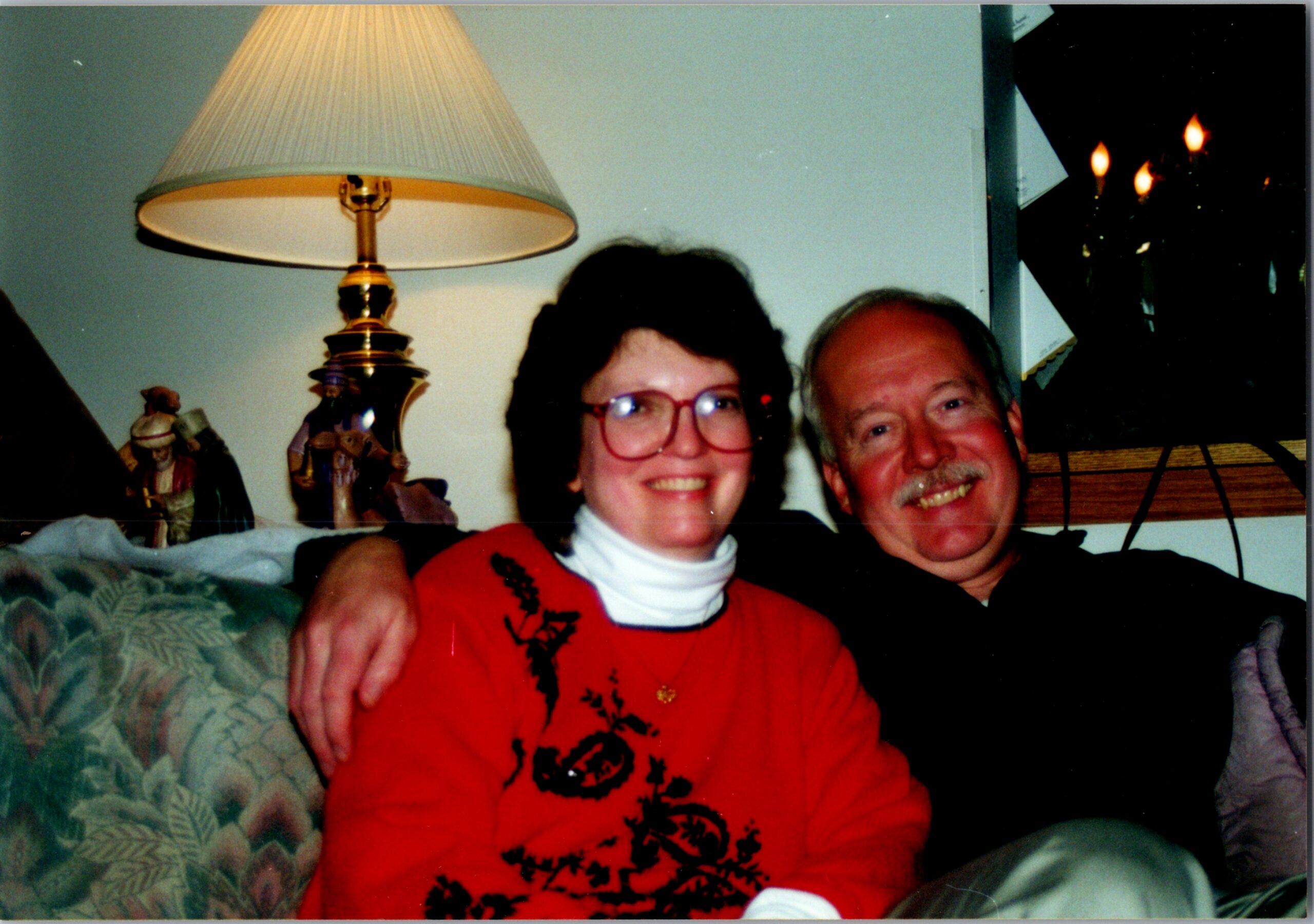 Two smiling adults sit close together on a couch in a cozy living room, with a lamp and figurines in the background.