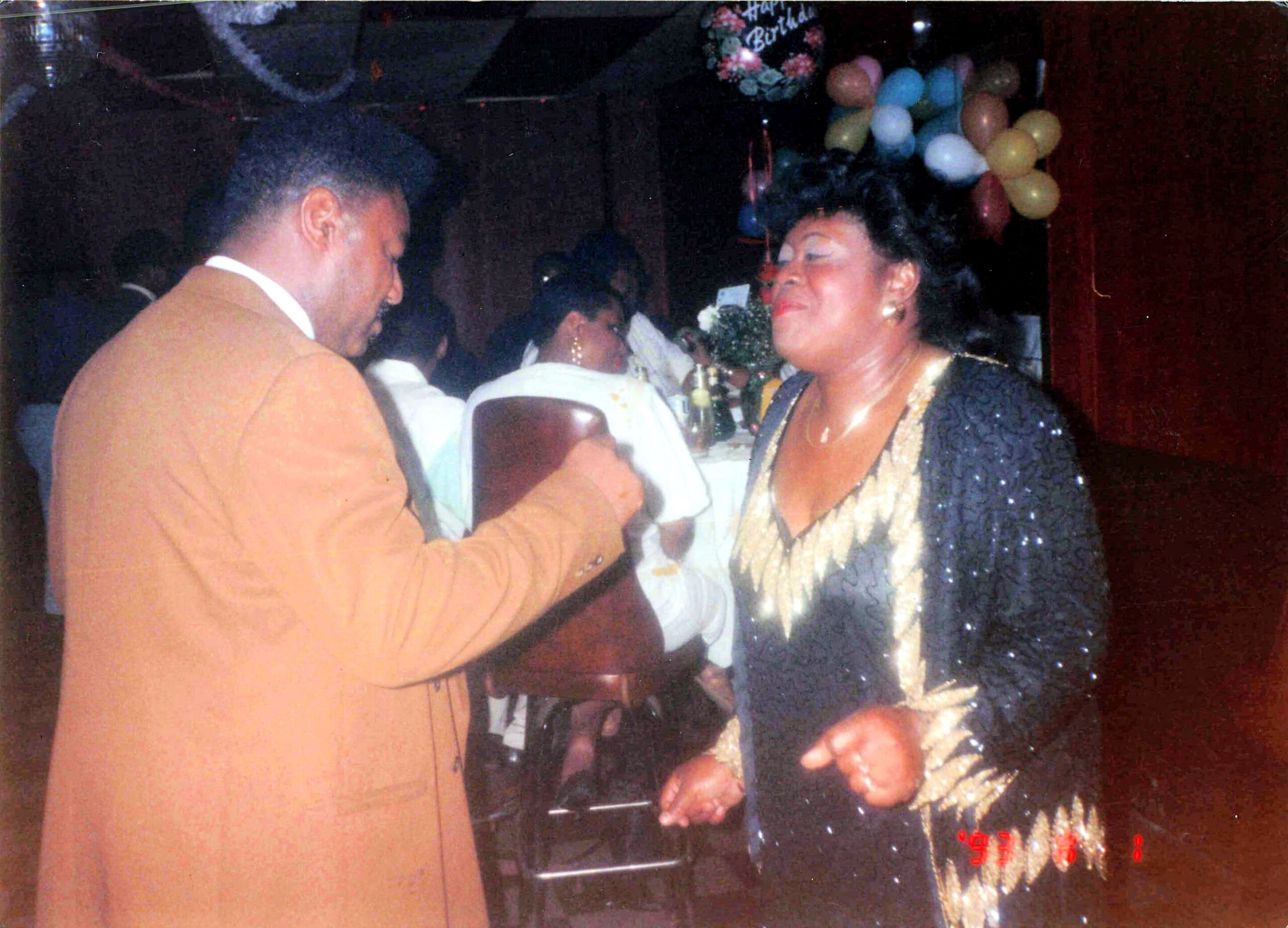 Couple dancing at a party, woman in a sequined dress and man in a tan suit, festive balloons in background.