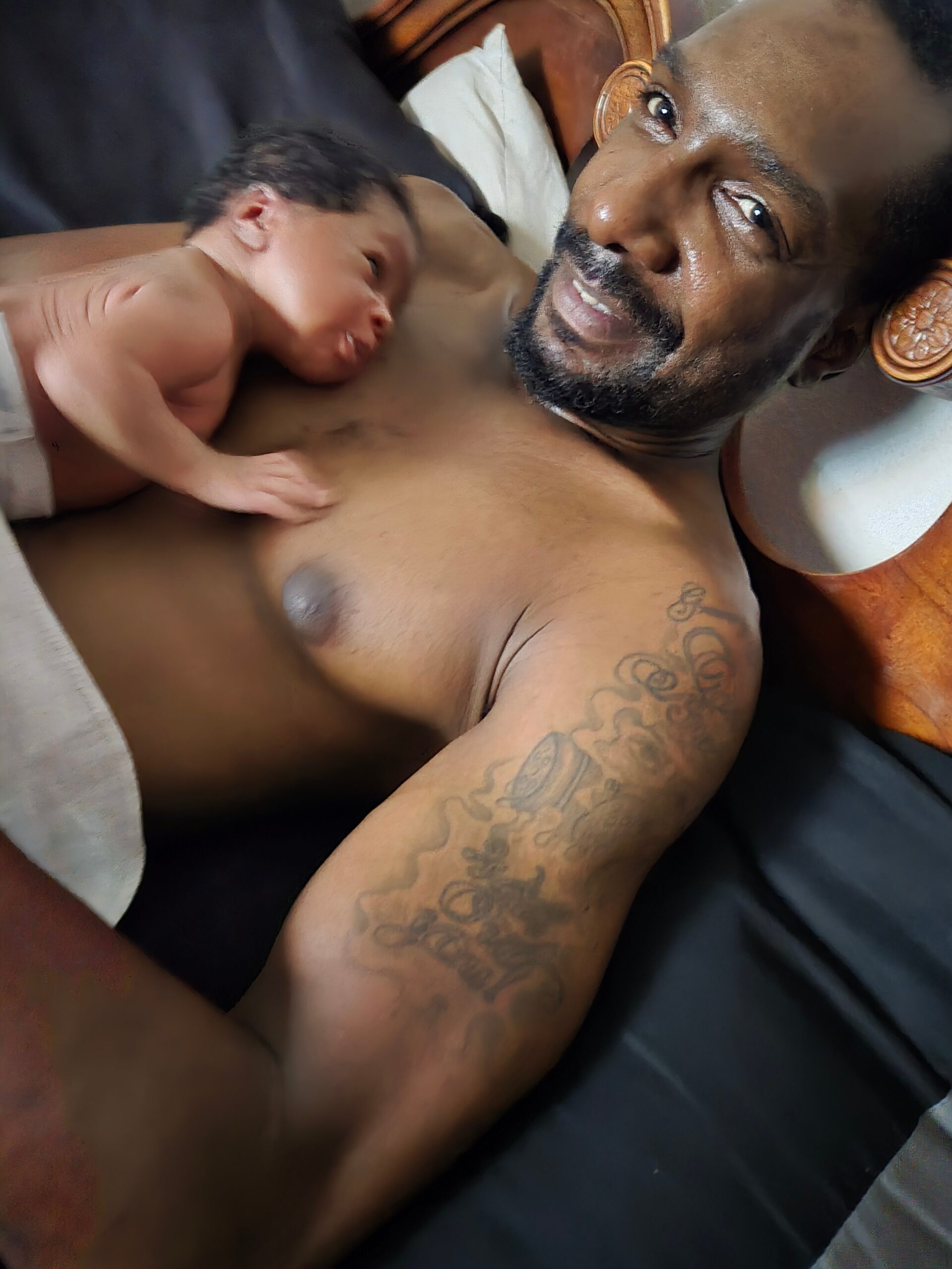 Father lying in bed with a newborn baby resting on his bare chest in skin-to-skin contact.