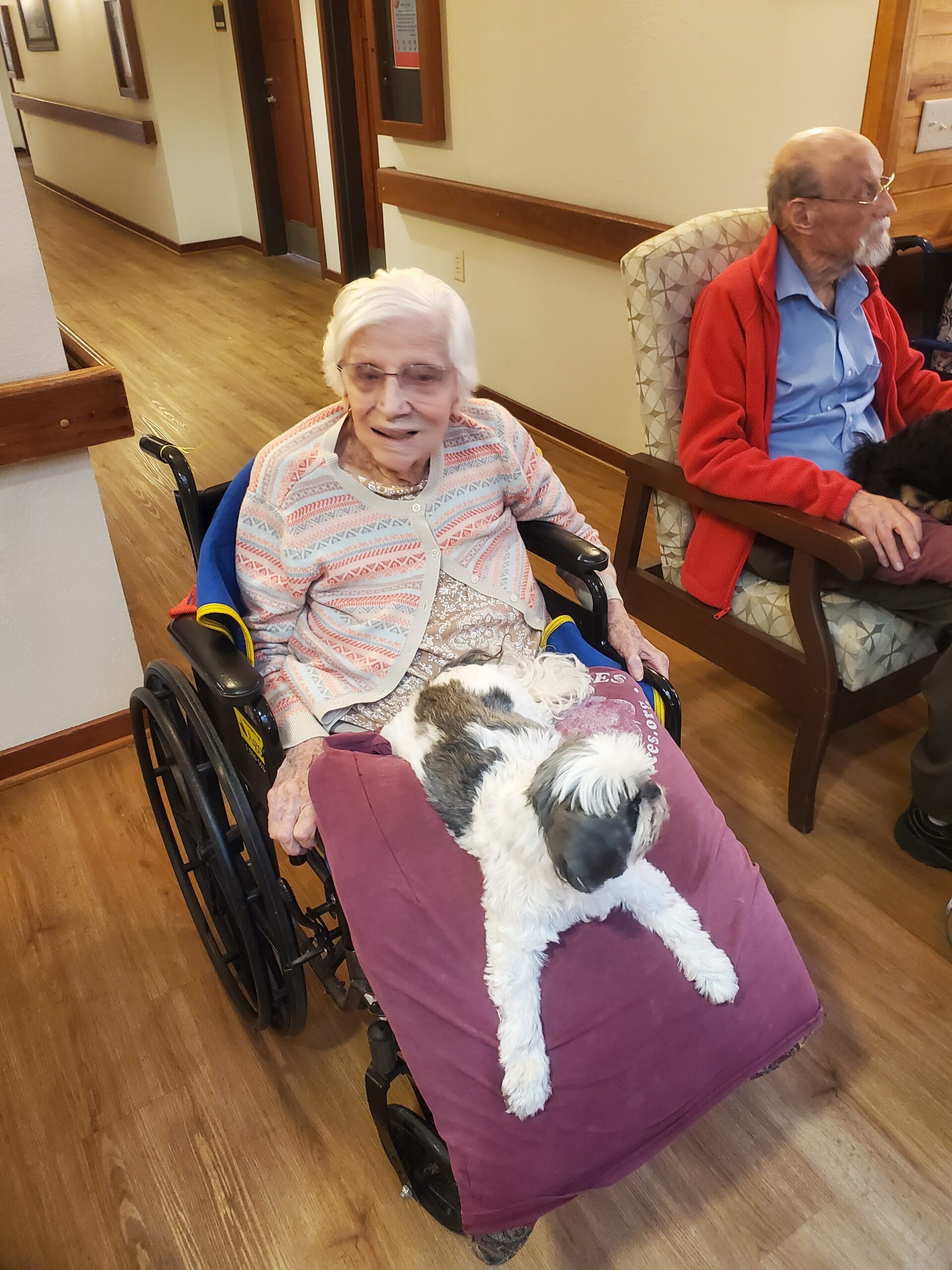 Elderly woman in a wheelchair with a small white-and-black dog on her lap in a care-home hallway; man in a red jacket sits nearby.