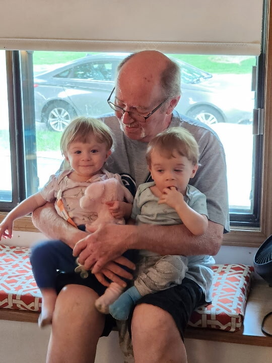Grandparent sitting on a bench by a window, holding two small children who smile at the camera.