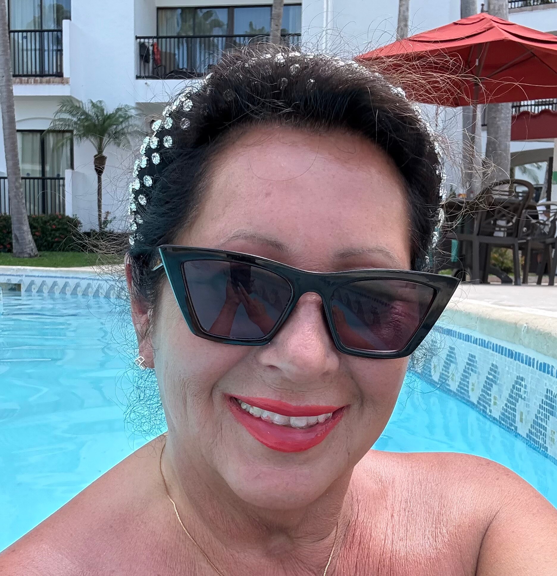 Close-up portrait of a woman by a pool wearing dark sunglasses and a pearl headband, smiling at the camera with red lipstick.