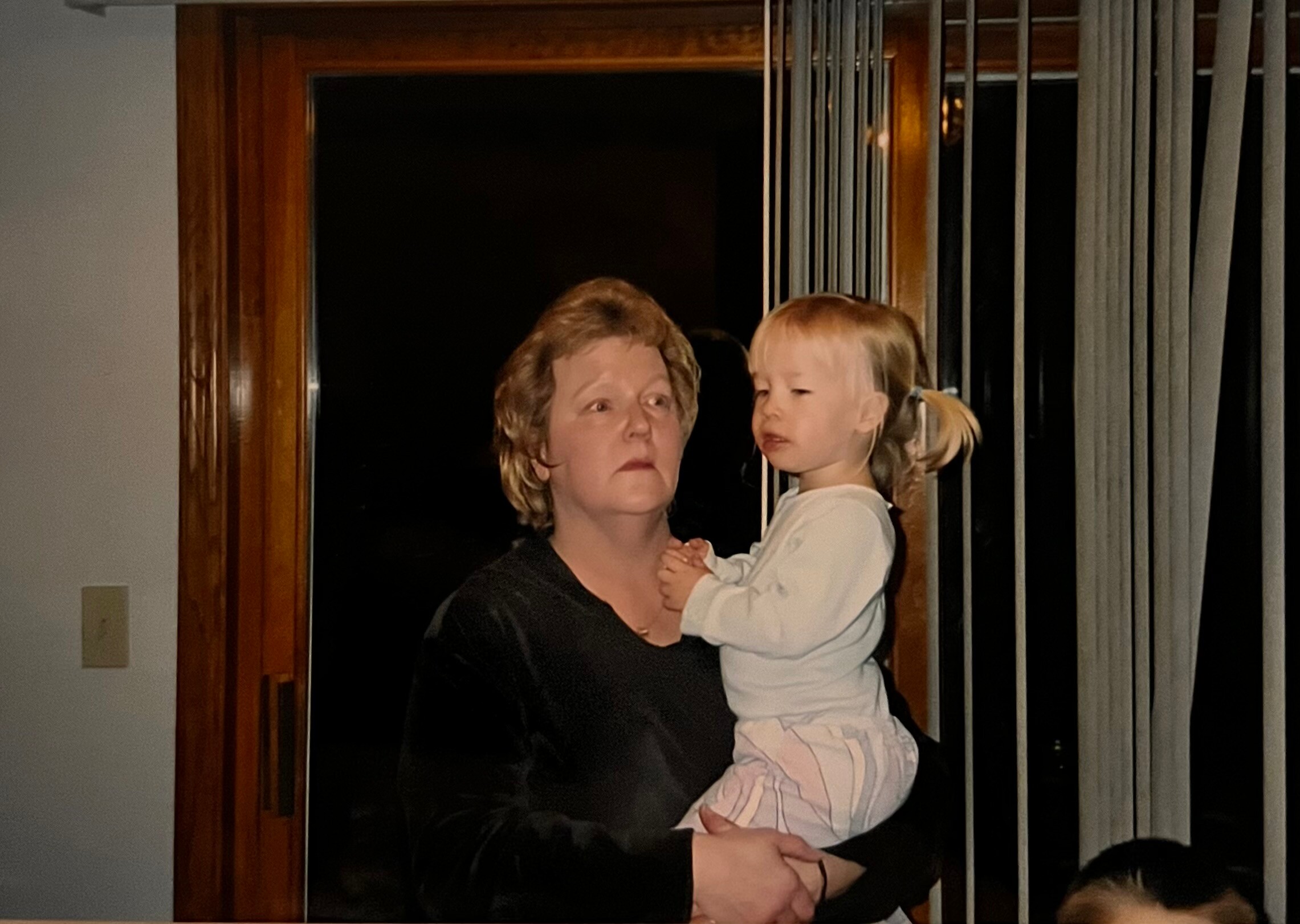 Woman holding a young child indoors near vertical blinds, facing left.