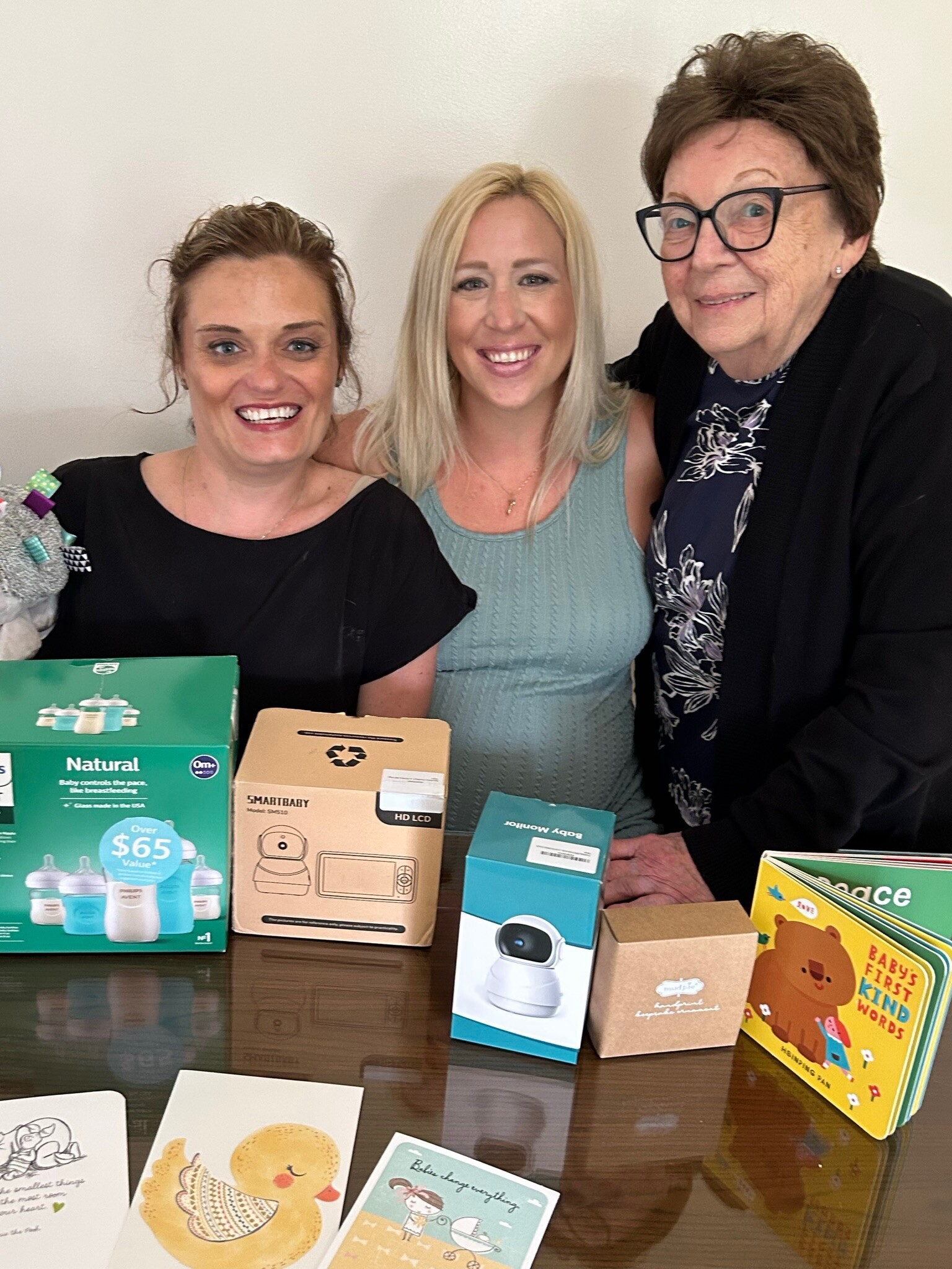 Three women smile at the camera behind a table with baby product boxes and cards on display.