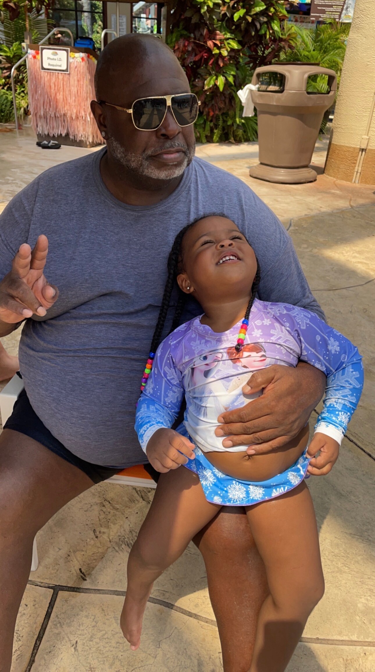Man wearing sunglasses and a blue T‑shirt sits outdoors while holding a smiling young girl in a purple Elsa-themed swimsuit; sunny day, tropical plants in background.