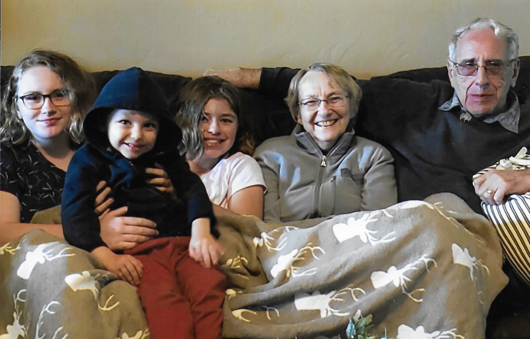 Family of five seated on a couch indoors, smiling at the camera with a floral throw draped over their laps.