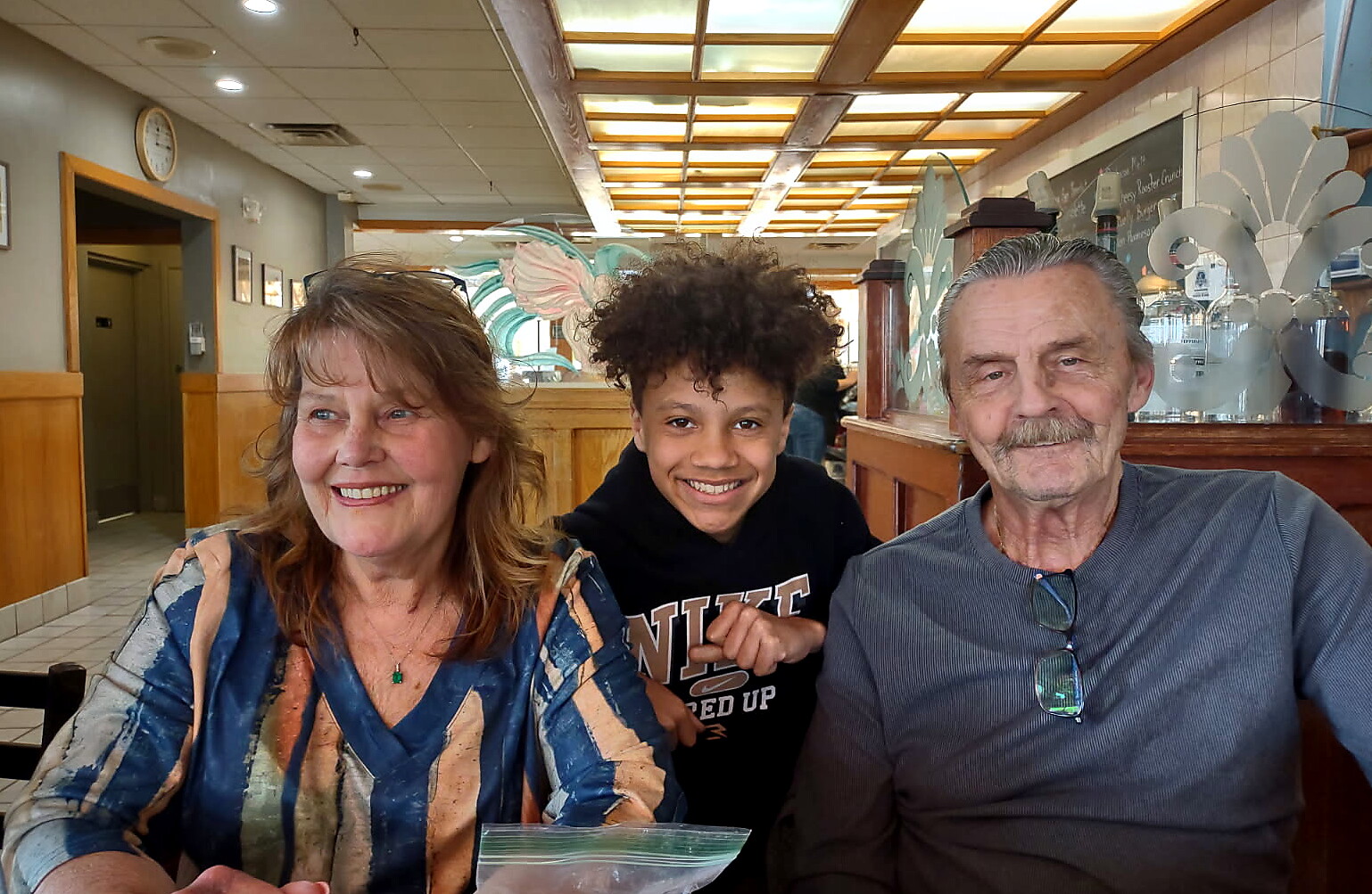 Three generations of family sit together at a booth in a bright cafe, smiling at the camera.