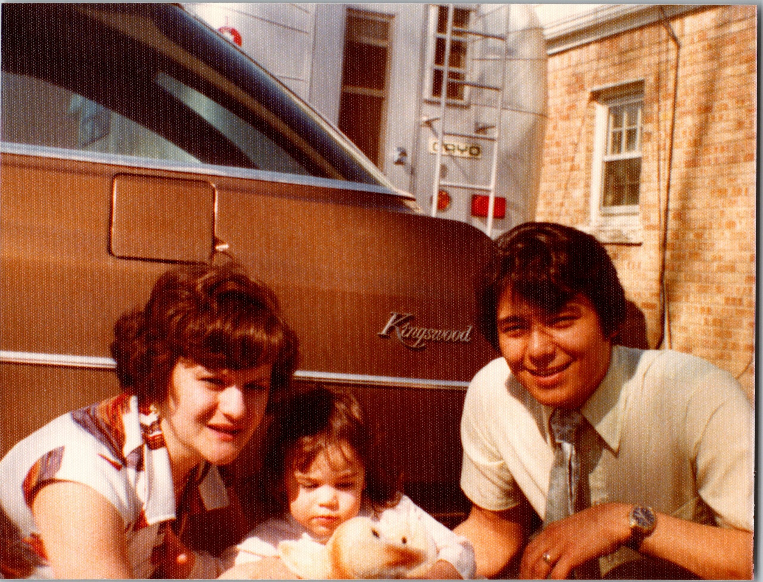 A smiling family group of five posing closely outside a brick house beside a brown vintage van; the van shows the word ‘Kingswood’ on its side.