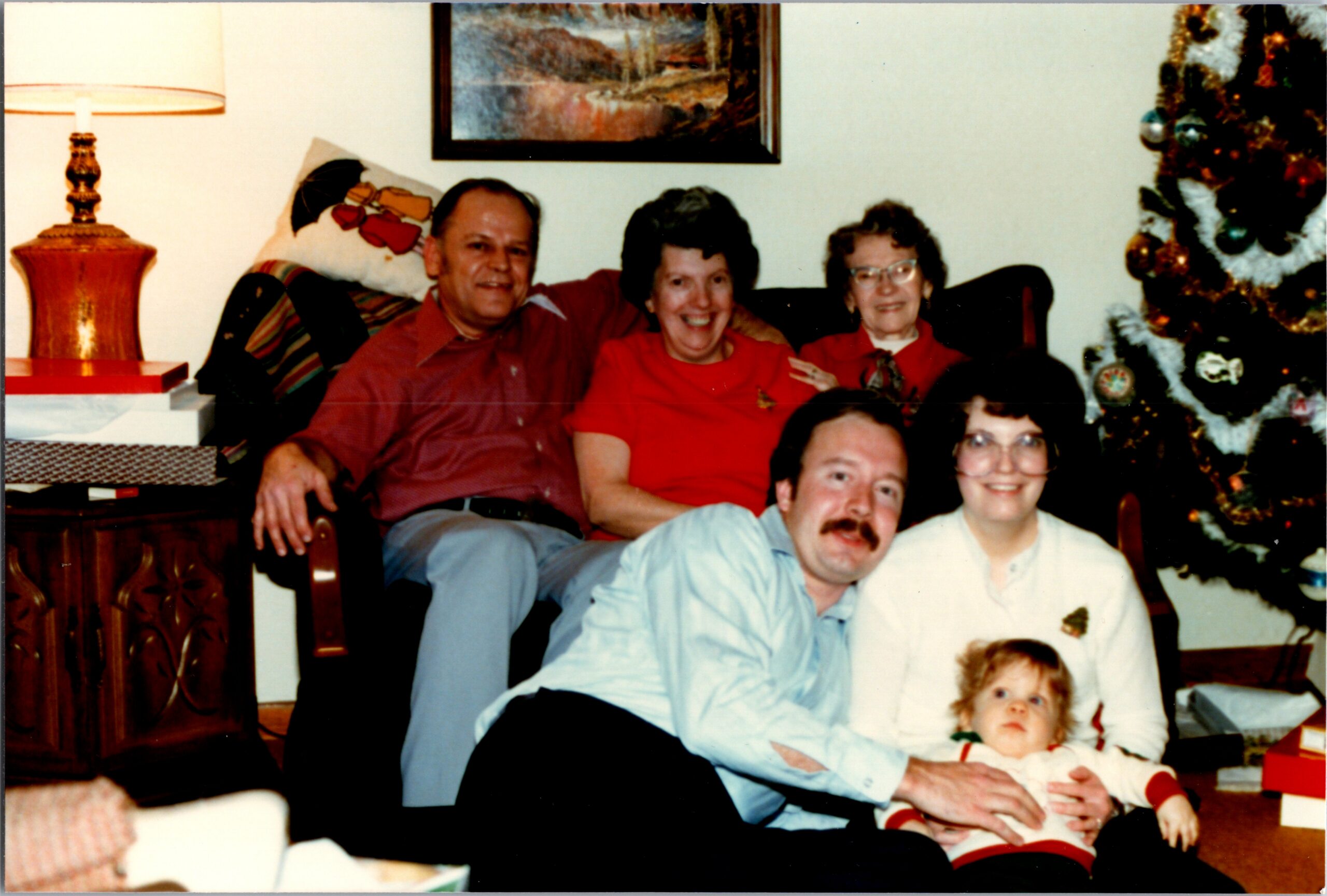A smiling family sits on a sofa in a cozy living room during Christmas, with a baby in front and a decorated tree to the right.