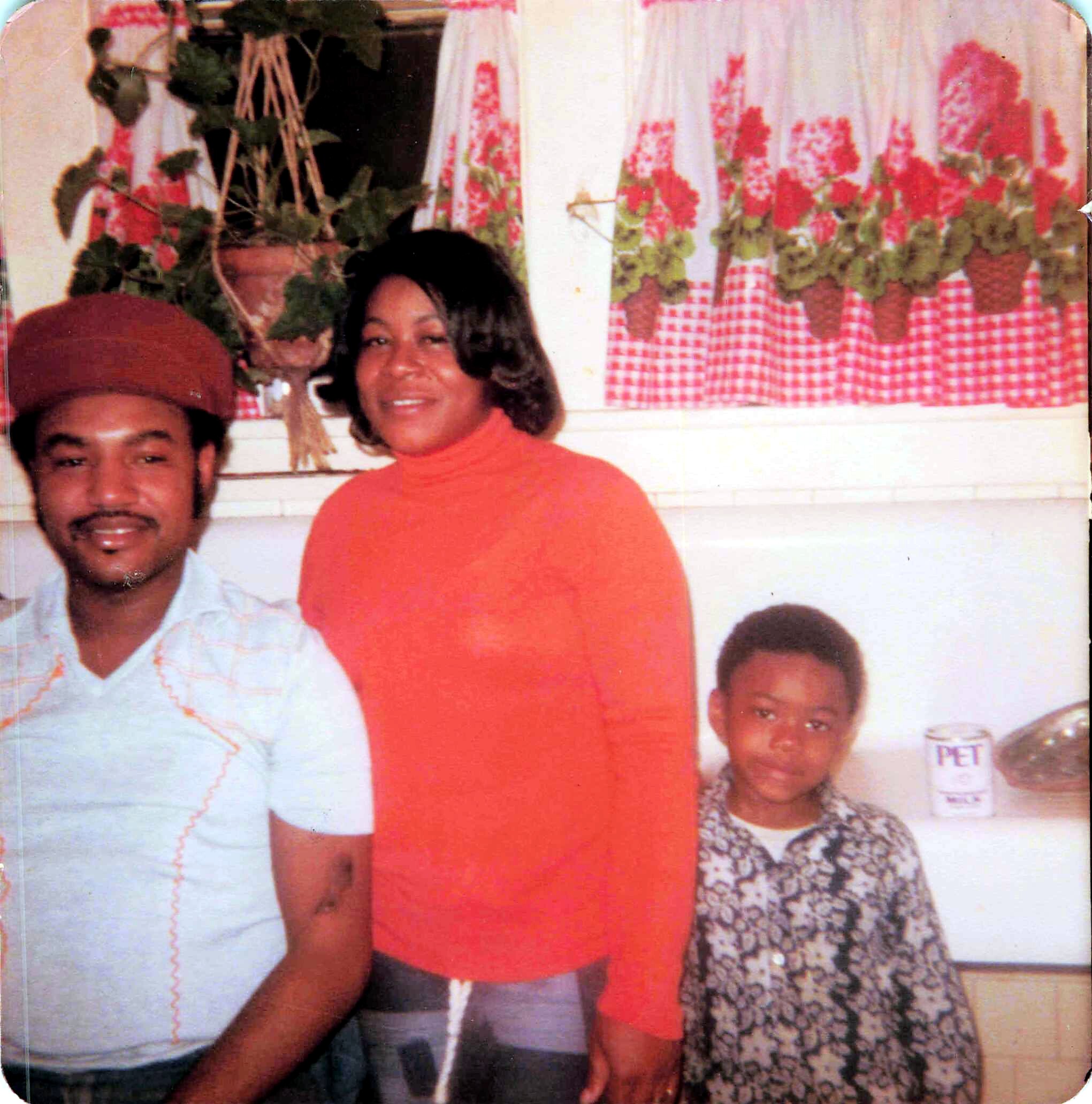 Woman in an orange top stands beside a man in a white shirt at a festive gathering, with red‑and‑white checkered tablecloths and potted flowers in the background.