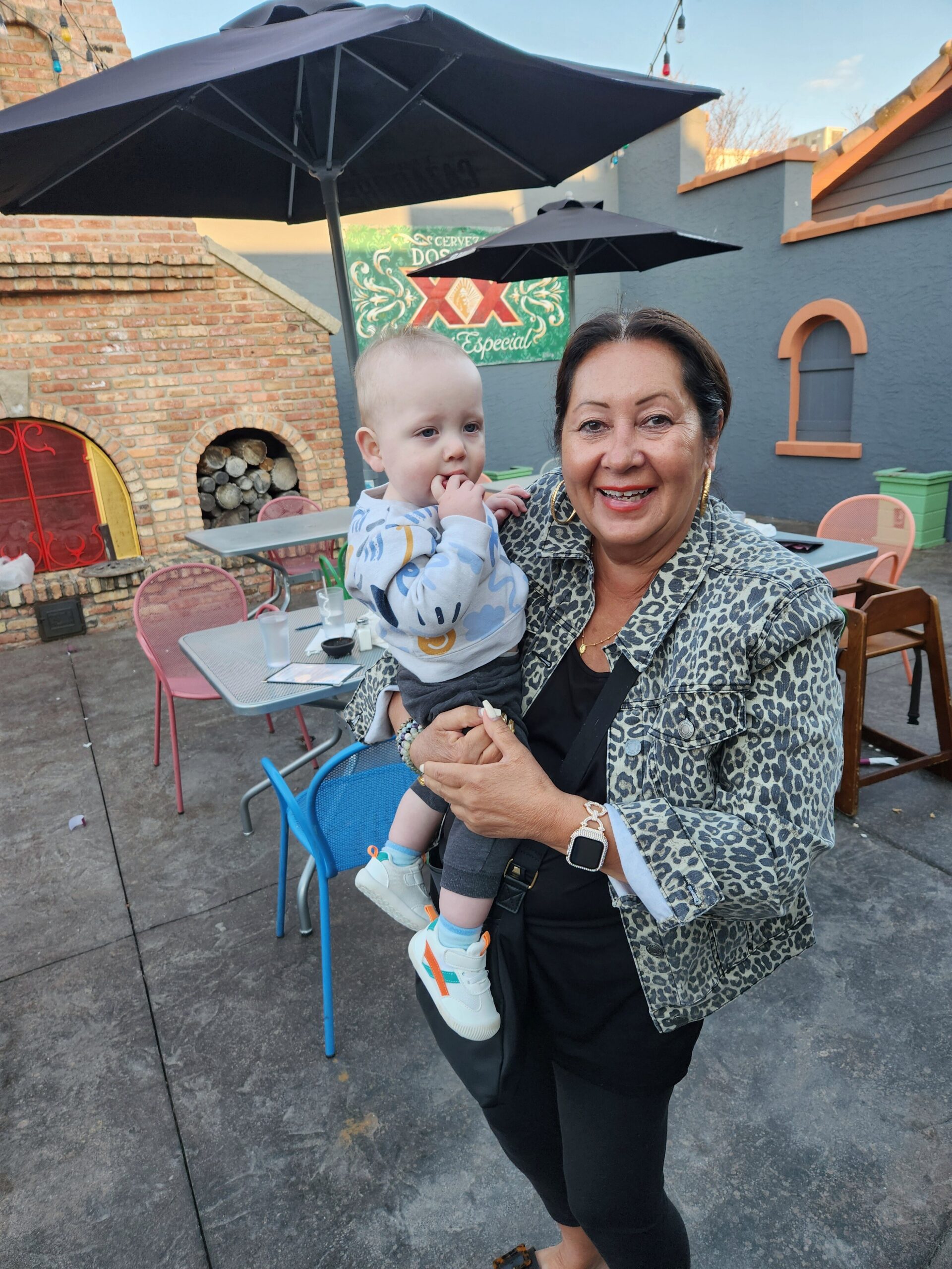 Smiling woman in a leopard-print jacket holding a baby outdoors at a cafe with brick oven and tables under umbrellas.