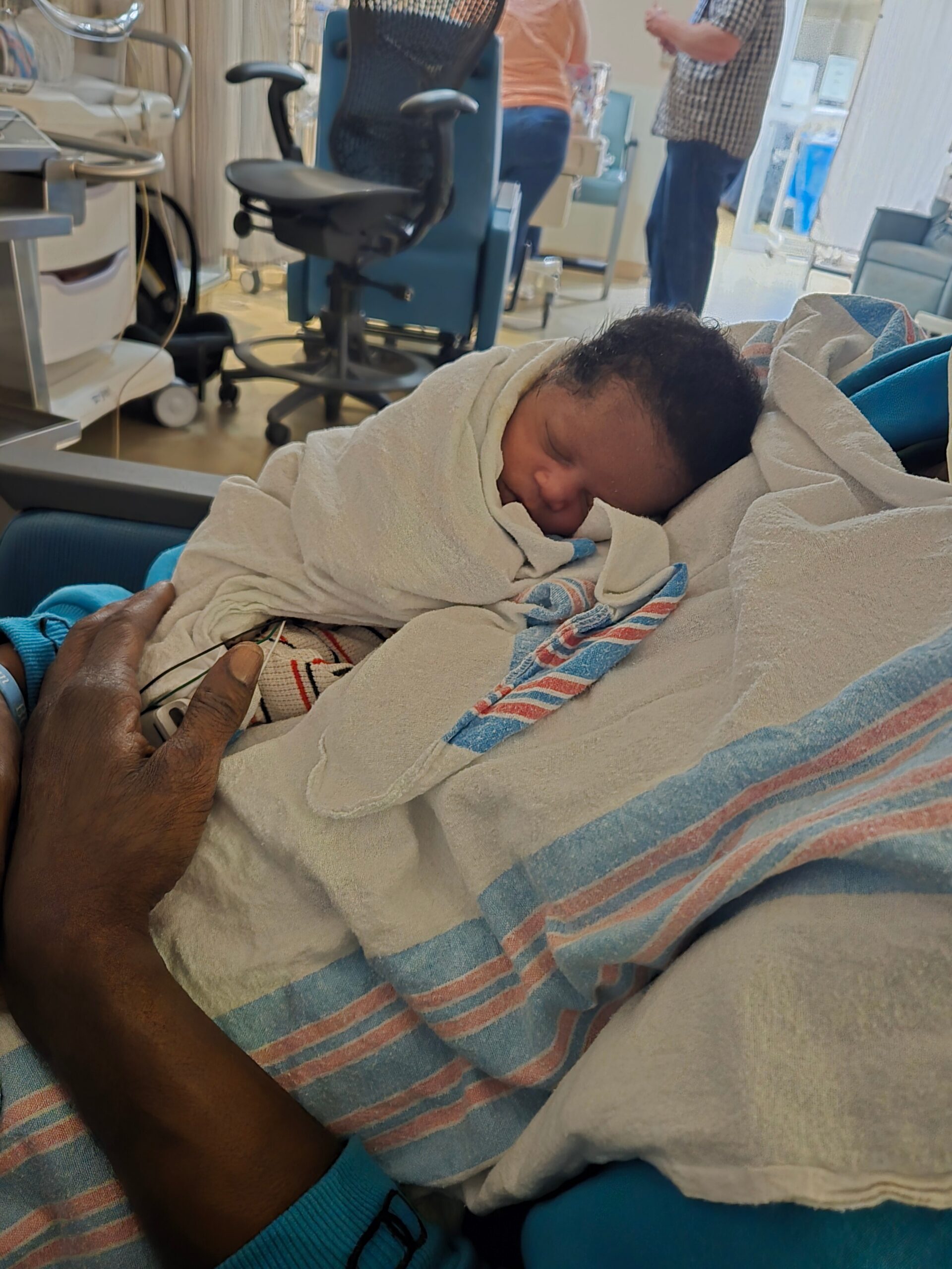 Newborn baby swaddled in blankets resting on a caregiver's lap in a hospital room with medical equipment in the background