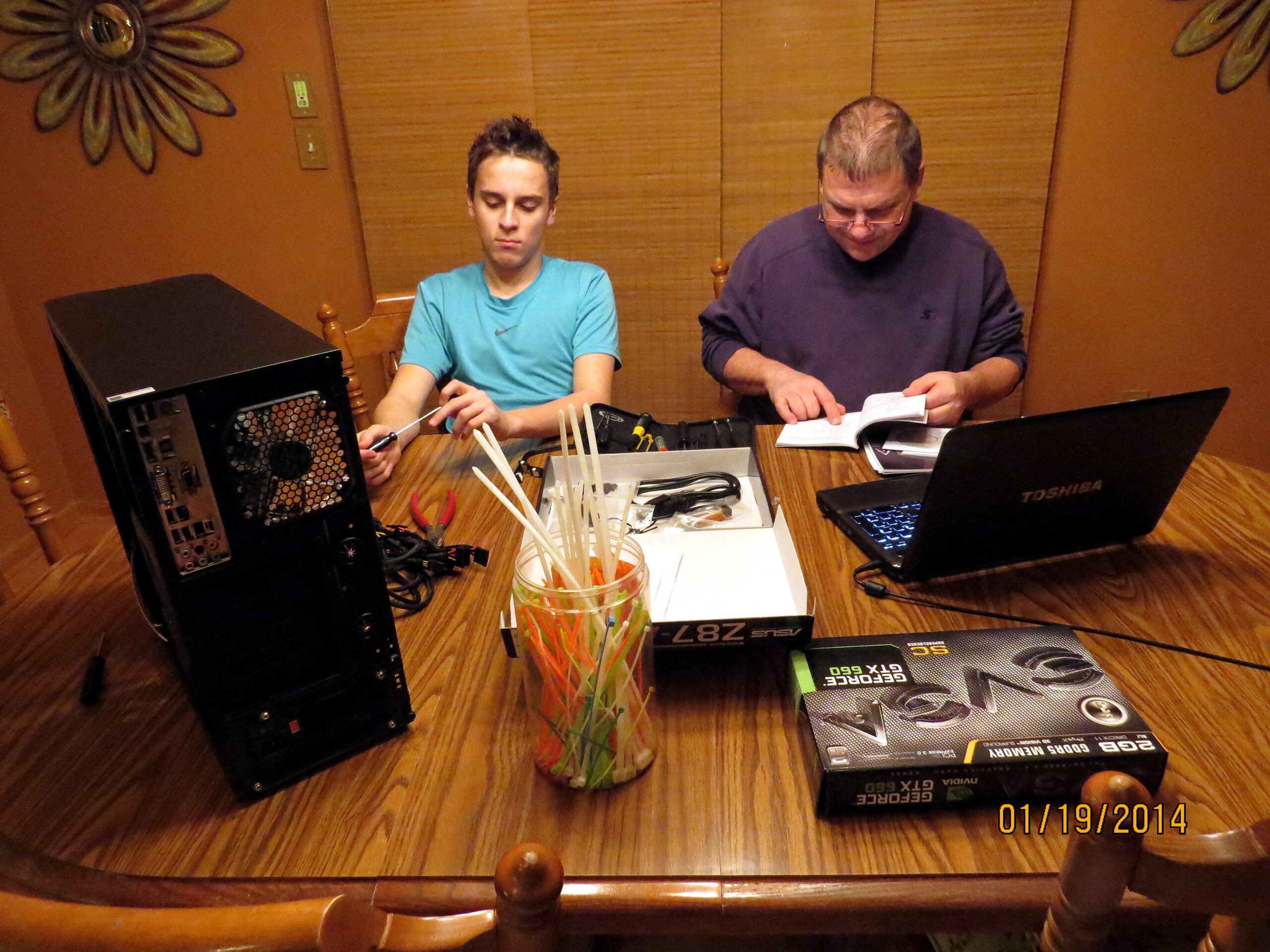 Two men sit at a wooden table assembling computer components; parts, tools, and a laptop are spread out, with graphics card boxes nearby.