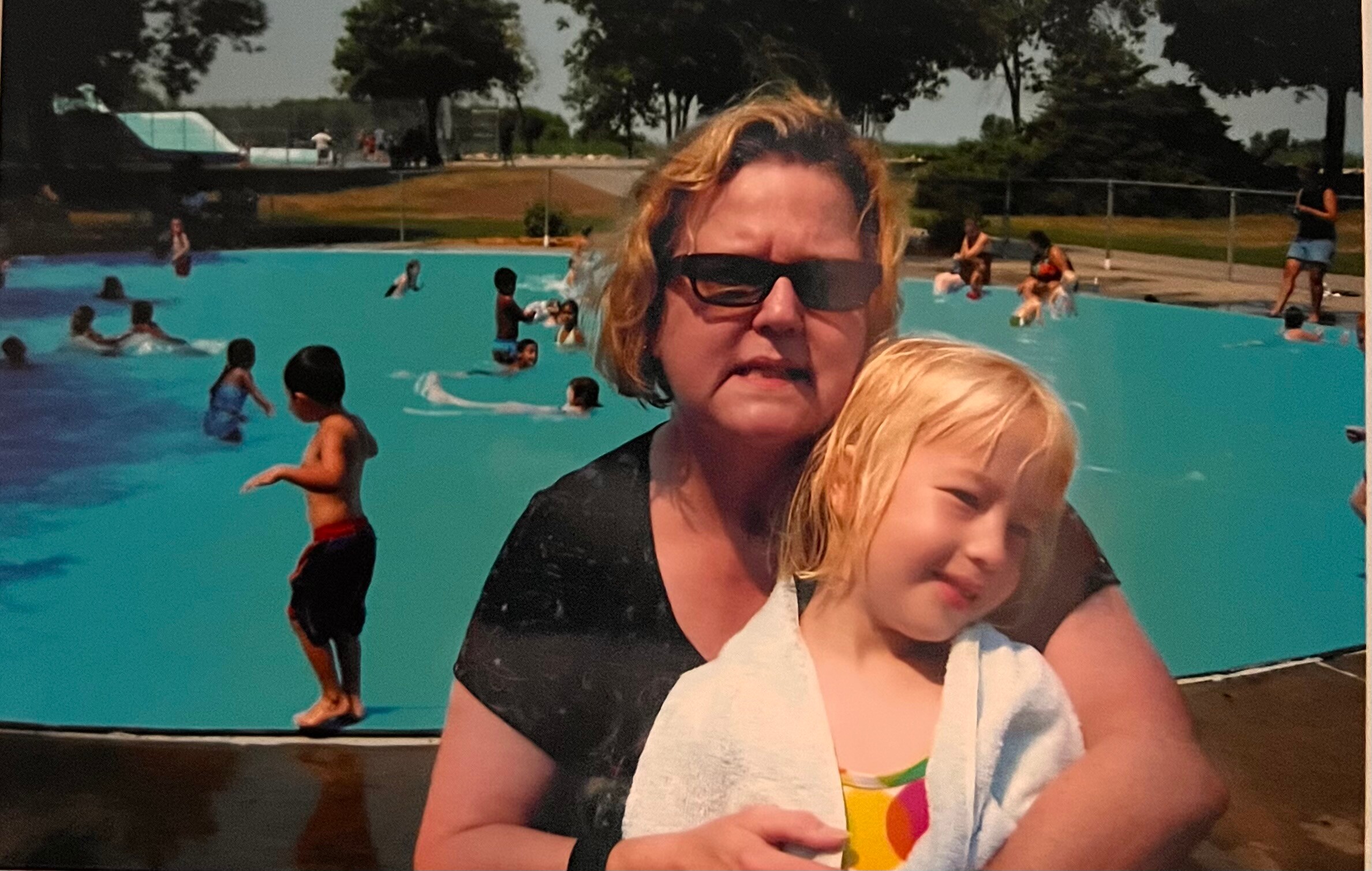 Woman wearing sunglasses holds a smiling child in front of a bright blue pool with people swimming in the background.