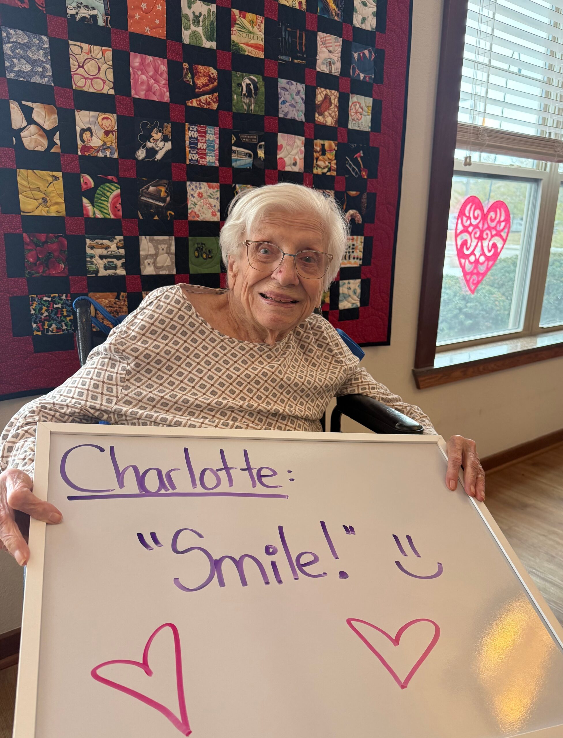An elderly woman in a patterned hospital gown sits in a wheelchair, holding a whiteboard that reads Charlotte: 'Smile!' with purple handwriting and a drawn pink heart, in front of a patchwork quilt and a window with a pink heart sticker nearby.