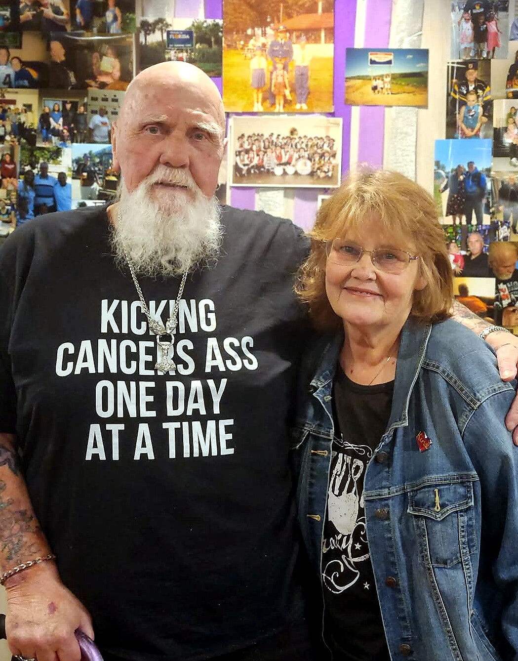Bearded man in a black shirt that reads 'KICKING CANCERS ASS ONE DAY AT A TIME' stands beside a smiling woman in a denim jacket in front of a wall covered with photos.