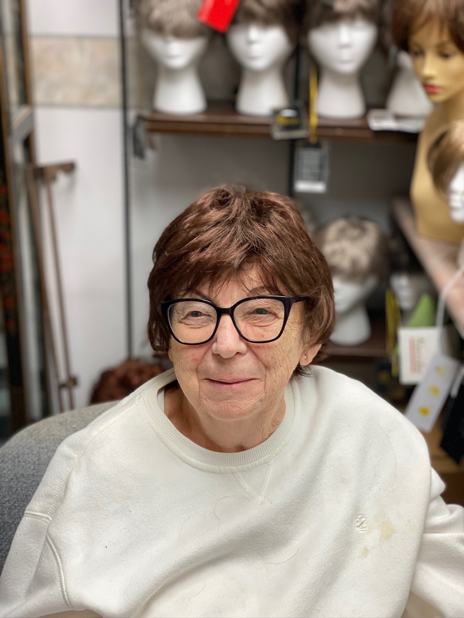 Close-up of an elderly woman with short brown hair and black glasses in a wig shop, mannequins with wigs blurred in the background.