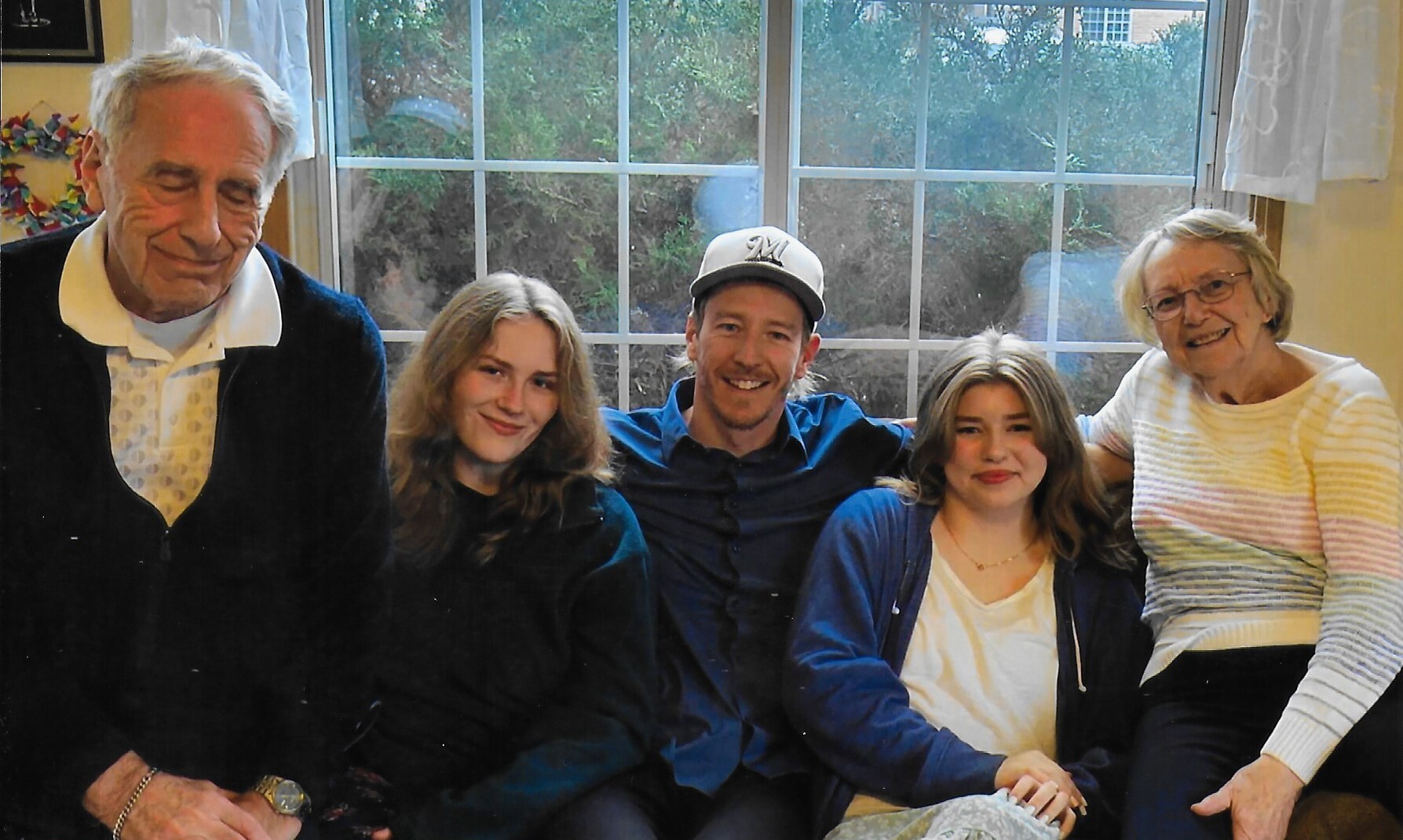 Five people sit closely together on a sofa in a sunlit living room, smiling at the camera with a window behind them.