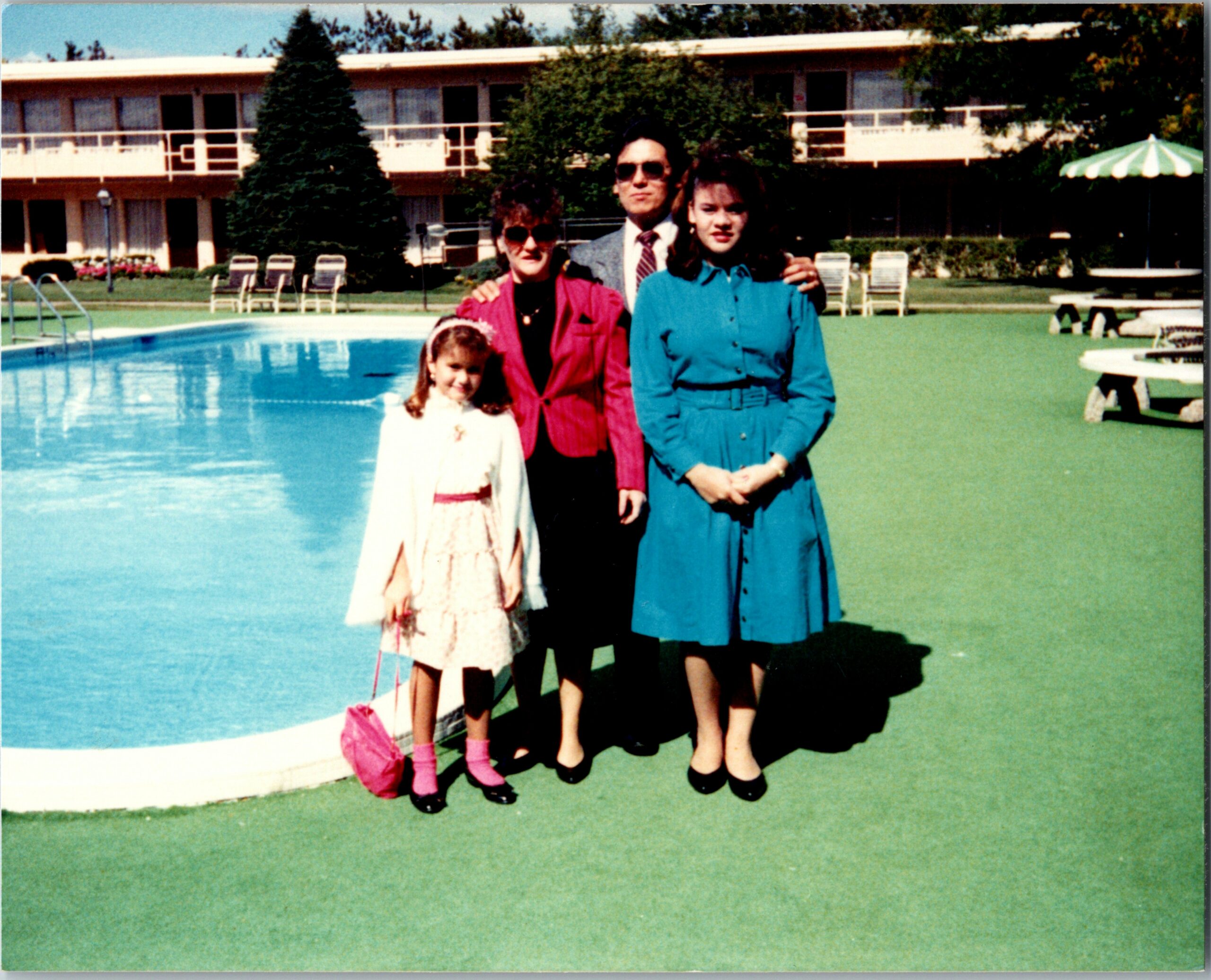 A family of four posing by a hotel pool: a girl in a white dress, two women in colored outfits, and a man in a suit, with lounge chairs in the background of a motel style building.