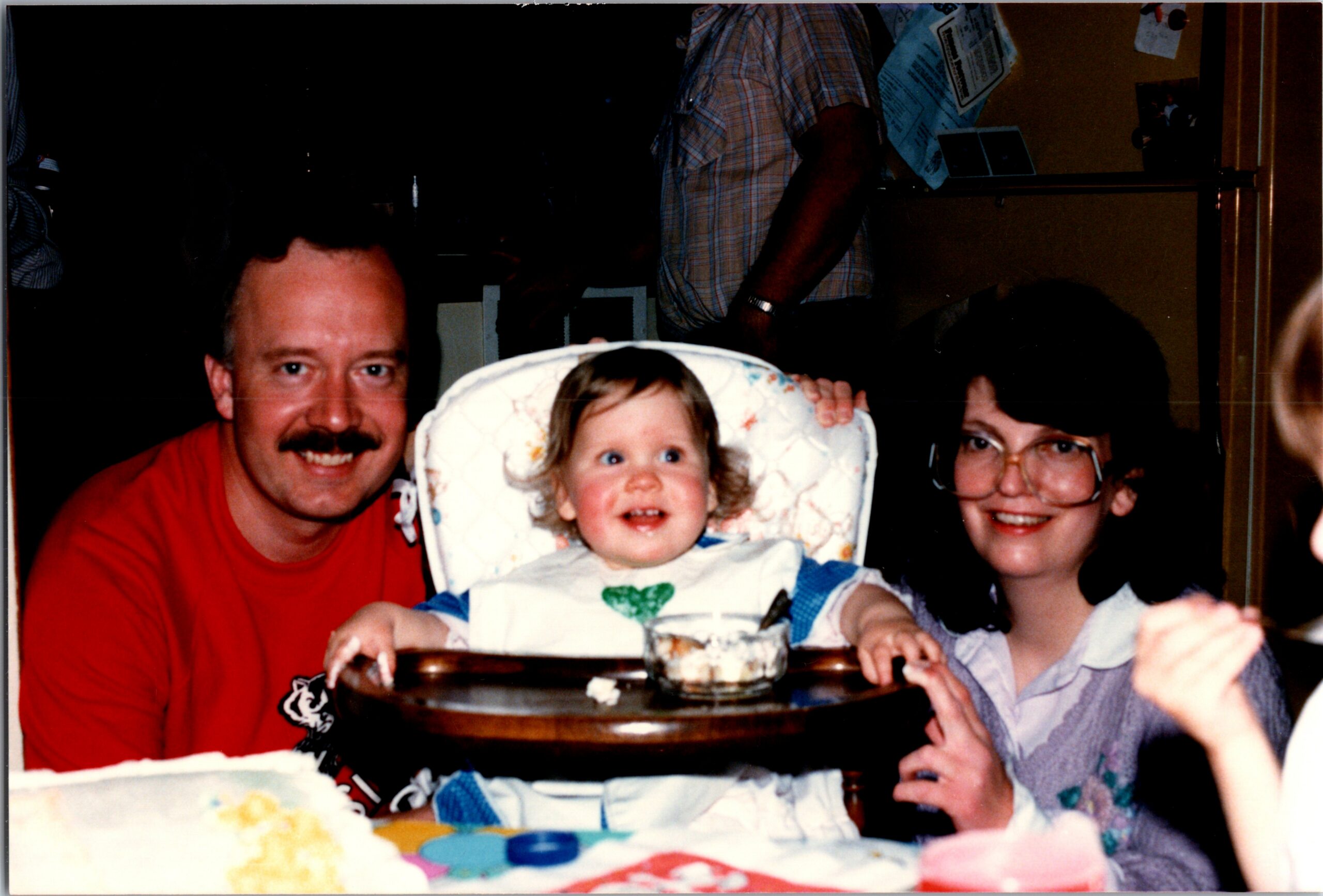 Man in a red shirt and a woman with glasses smile beside a baby in a high chair at a birthday table with cake.